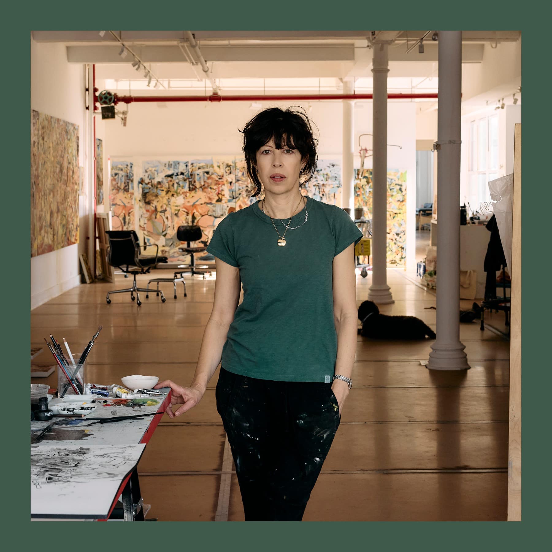 Cecily Brown in a green T-shirt and paint-spattered trousers standing in her New York studio, with her paintings behind her