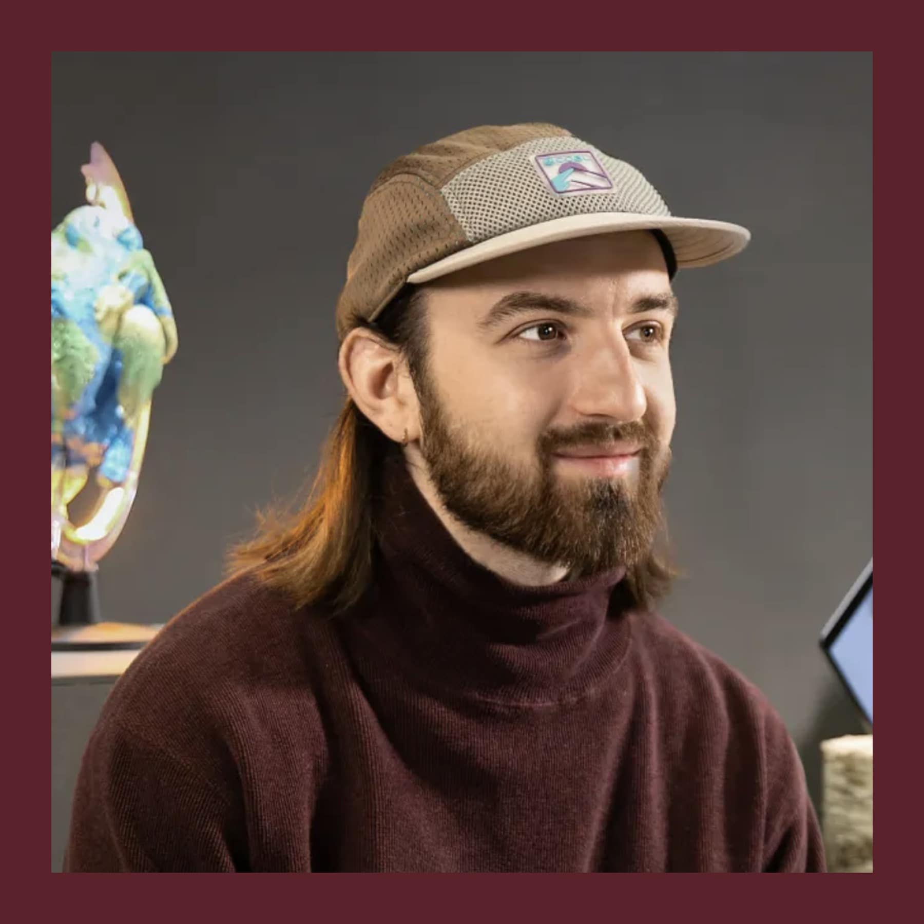 Delfino Fidel in a cap and a burgundy roll-neck jumper, smiling, beside one of his iridescent sculptures