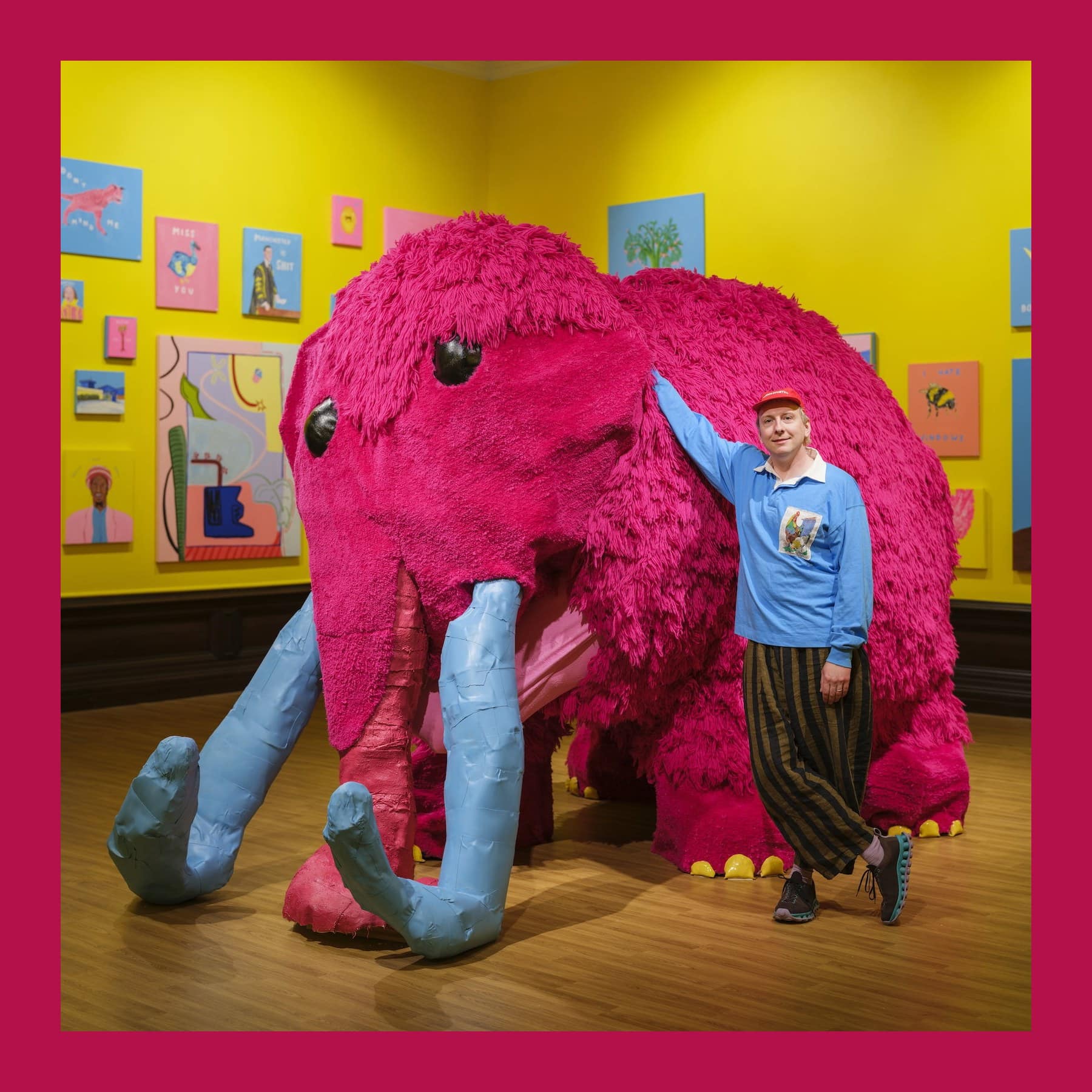 Joe Lycett leaning on his giant pink woolly mammoth sculpture in a yellow gallery hung with his paintings