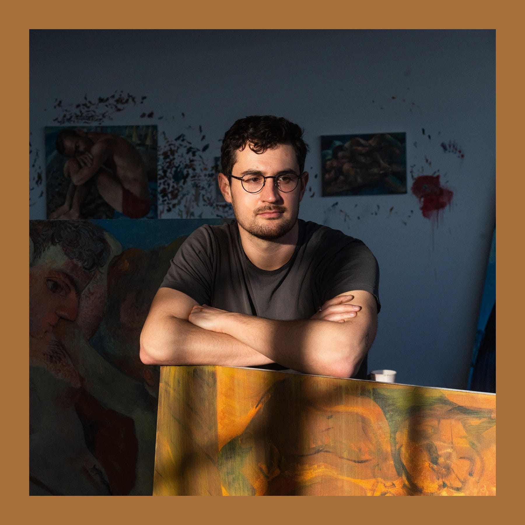 Louis Fratino with arms folded, leaning on a painting in his studio, with his figure paintings on the wall behind