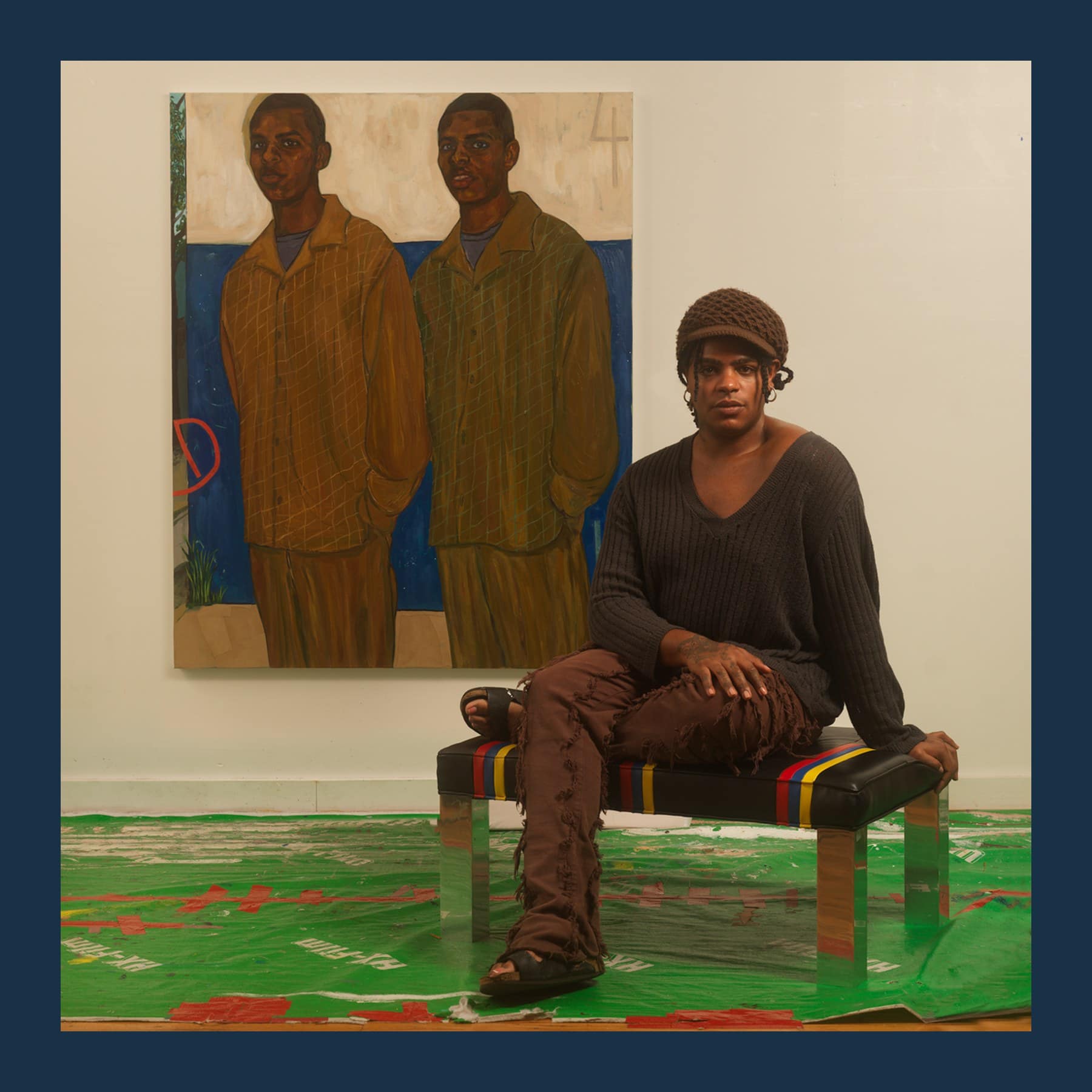 Khalif Tahir Thompson sitting on a striped bench in his studio in front of his painting of two young men