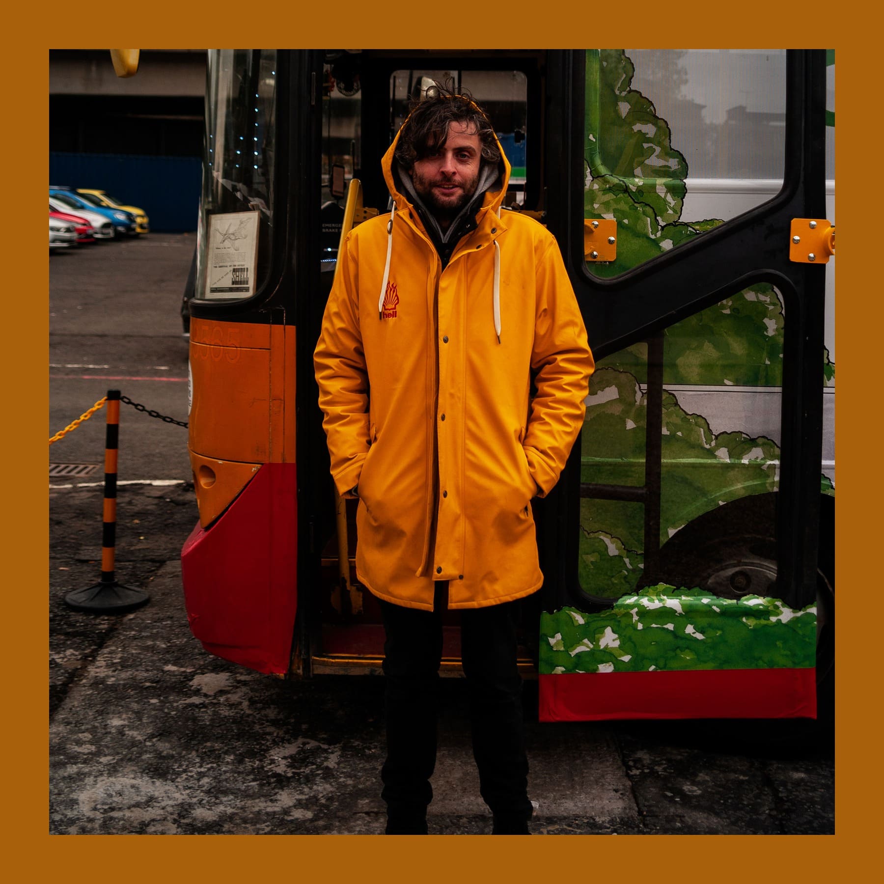 Darren Cullen in a yellow hooded raincoat with a Shell-style logo, standing in the doorway of his Hell Bus