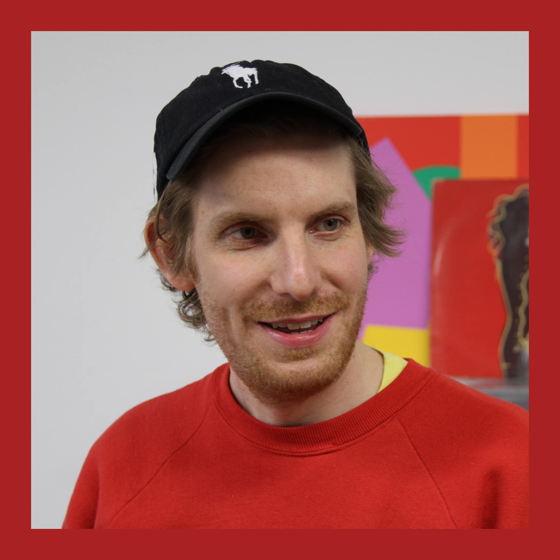 Cory Arcangel in a black cap and red sweatshirt, smiling in his studio in front of colourful works
