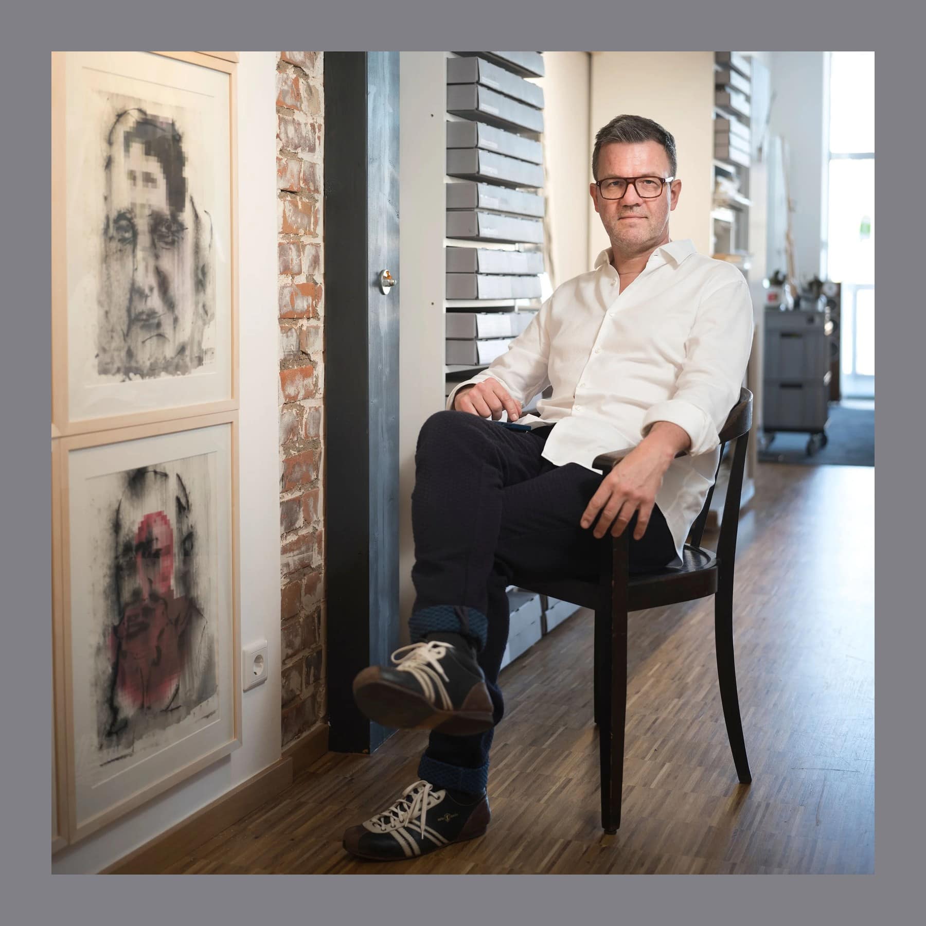 Jörg Gläscher in a white shirt and glasses, seated on a wooden chair in his Leipzig studio beside framed drawings
