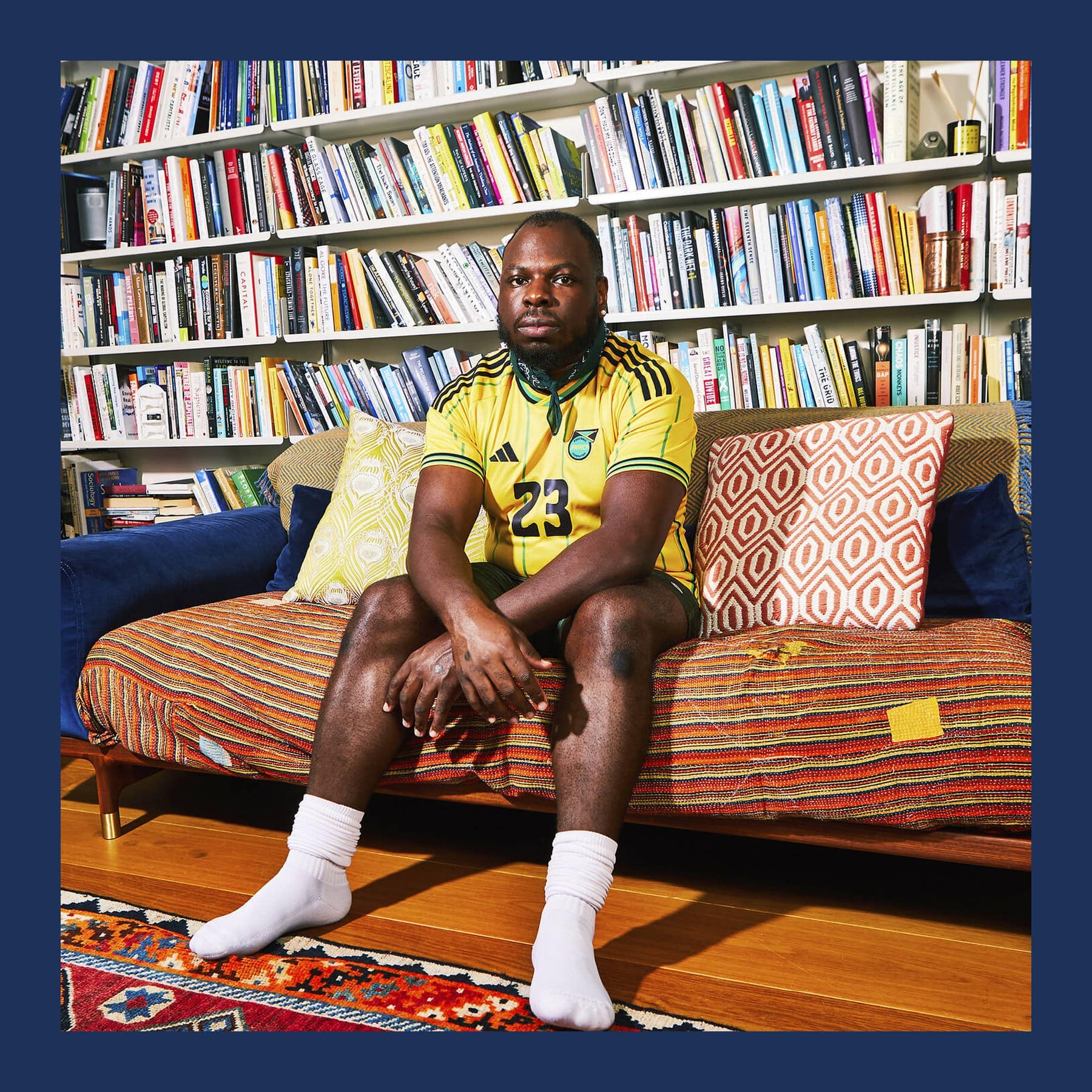 R.I.P. Germain in a yellow Jamaica football shirt sitting on a blue sofa in front of full bookshelves