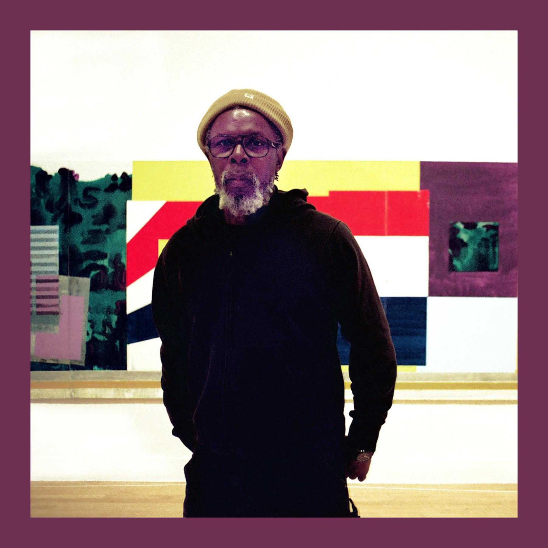 Hurvin Anderson in a mustard beanie and black hoodie, standing in front of one of his paintings at Tate Britain