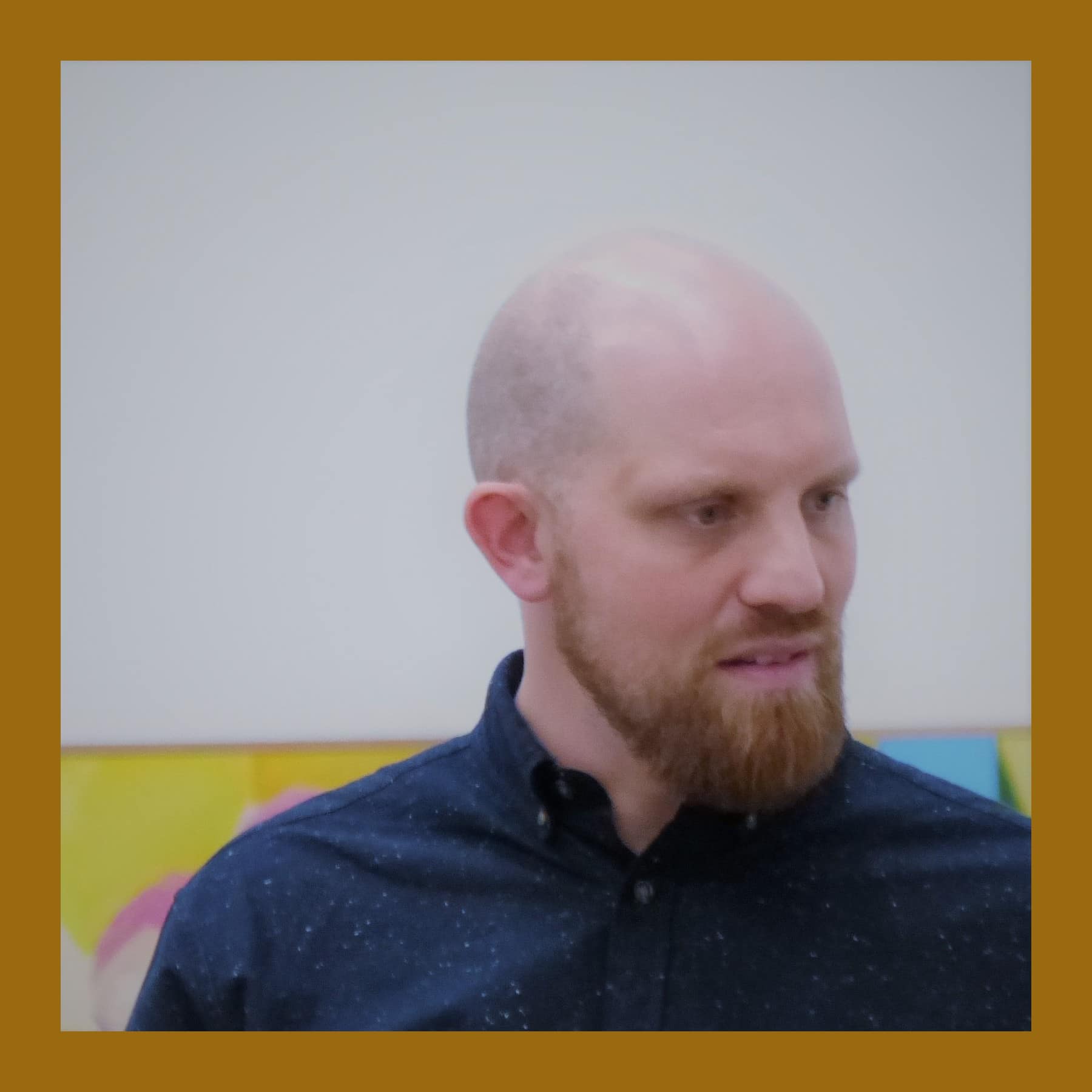 Ryan Mosley, bald and bearded in a dark shirt, in front of one of his colourful paintings