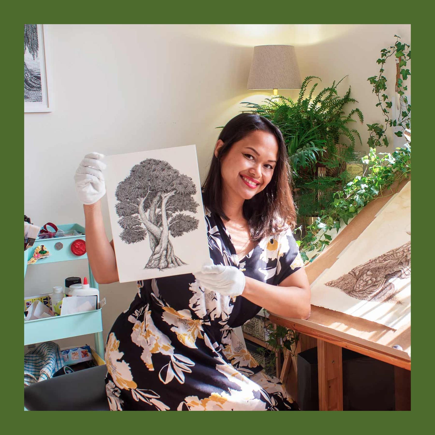 Salma Price-Nell at her drawing desk in white cotton gloves, holding up an ink drawing of a tree, with plants behind her
