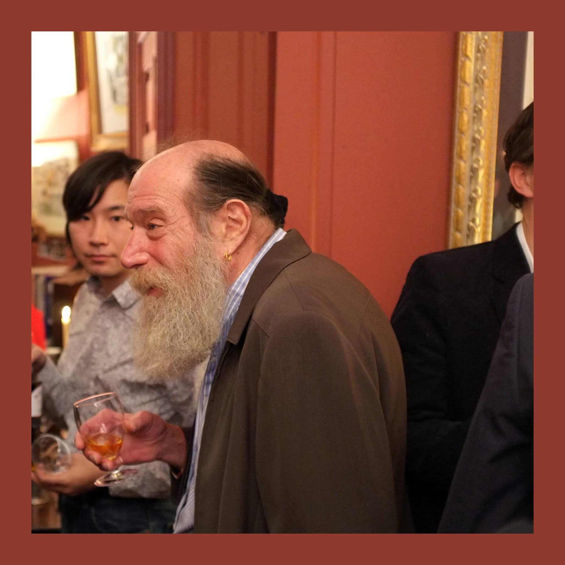 Lawrence Weiner, with a long white beard, holding a drink at a crowded reception