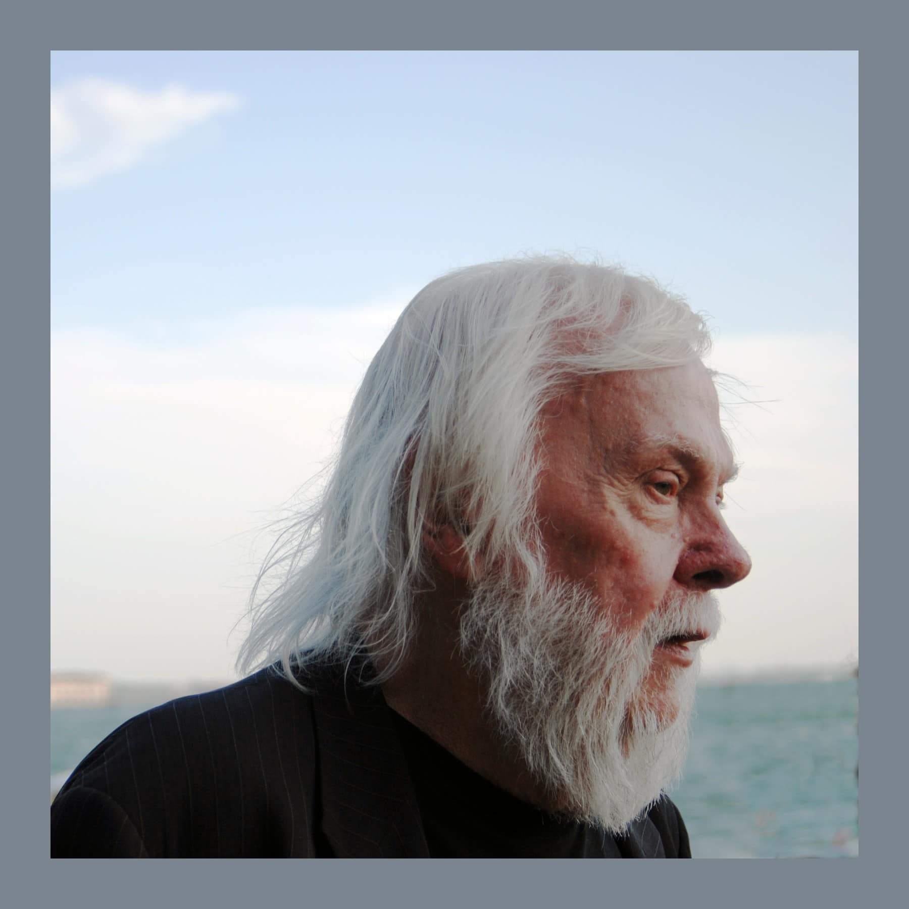 John Baldessari in profile with long white hair and a white beard, beside the lagoon in Venice