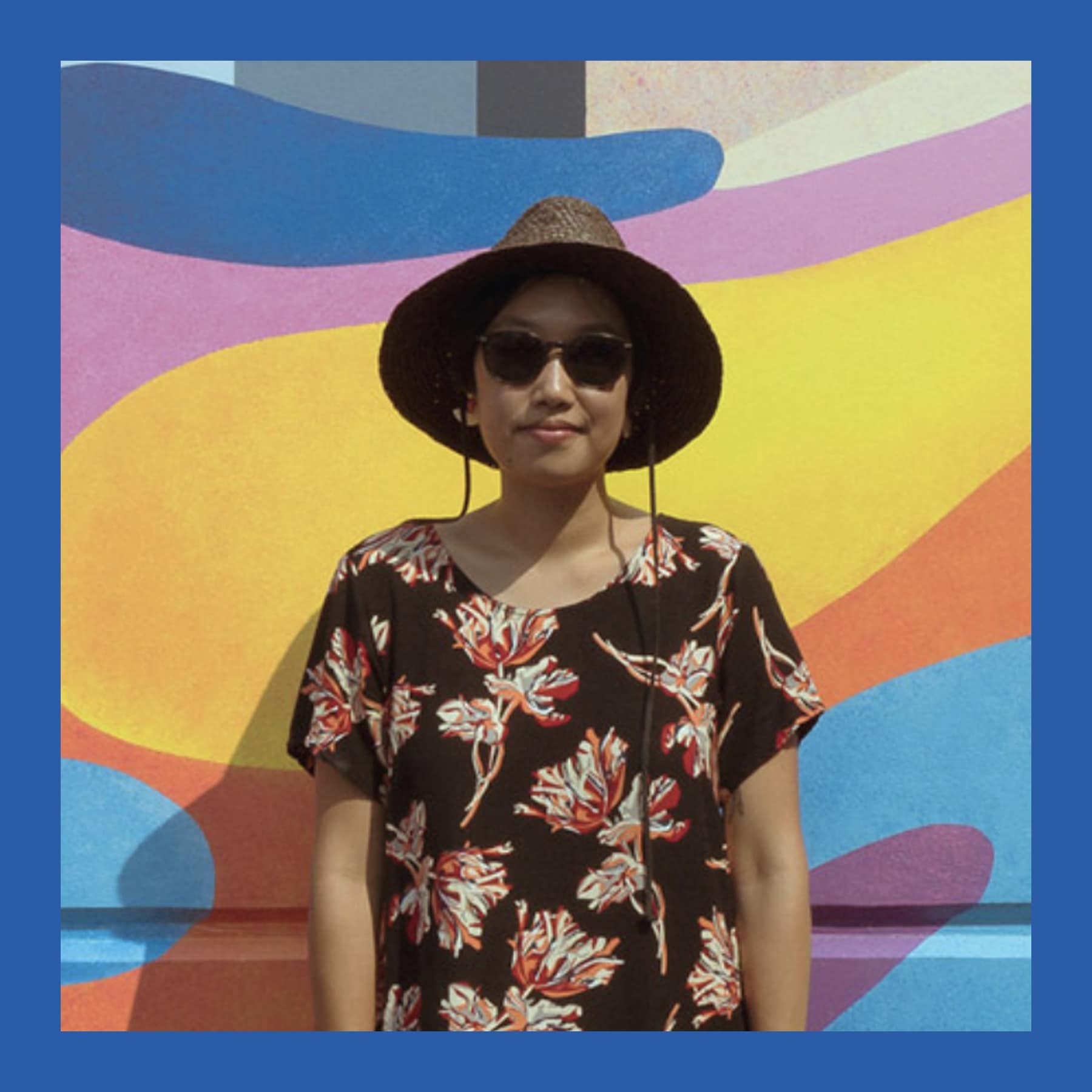 Hanna Lee Joshi in a sun hat, sunglasses and a floral dress, standing in front of a colourful mural