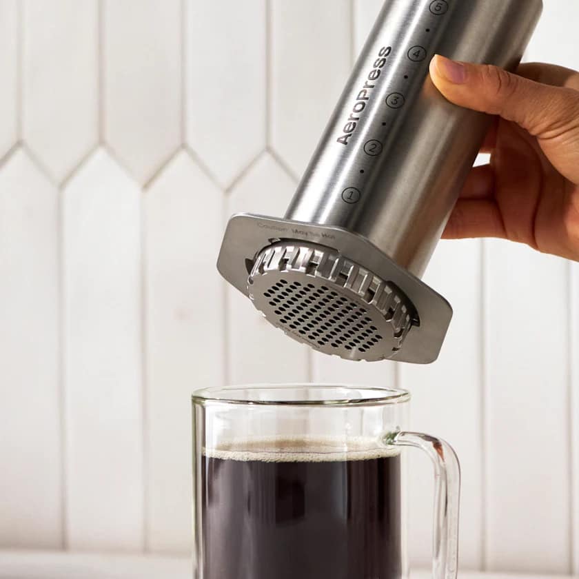 Hand pressing a stainless steel AeroPress Steel coffee maker into a clear glass mug of black coffee.