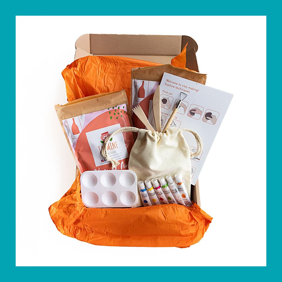 Open kraft box with orange tissue holding a clay pouch, wooden tools in a calico bag, six paint tubes, a white palette and a printed guide.