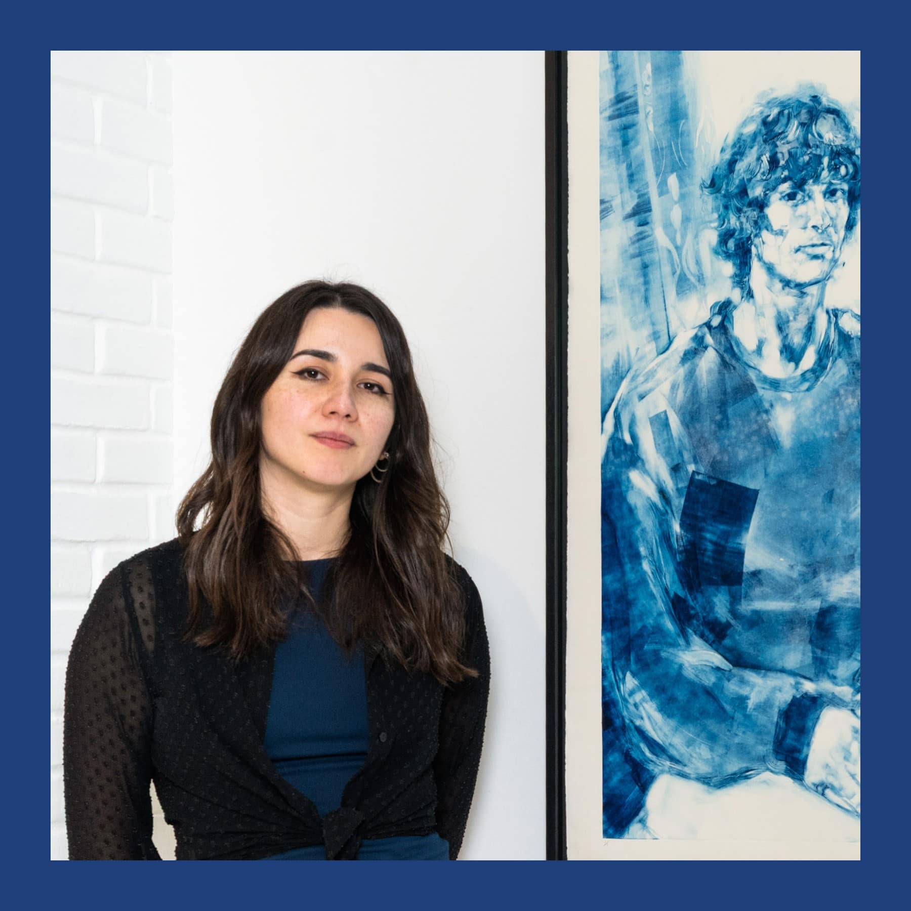 Chloe Barnes in a black cardigan standing in a white gallery beside one of her large blue monotype portraits