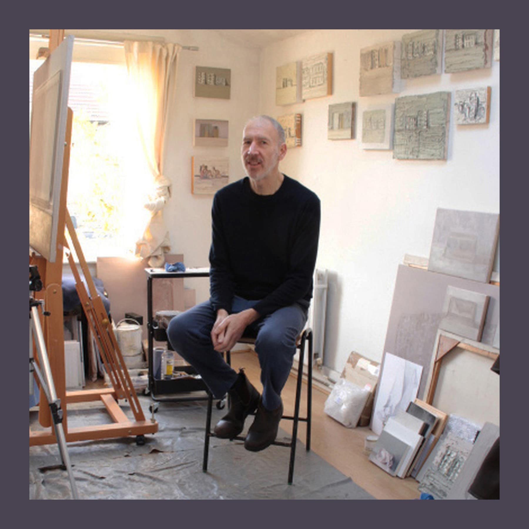 Alex Hanna sitting on a stool in his bright studio, with an easel to one side and small grey still-life paintings on the wall behind