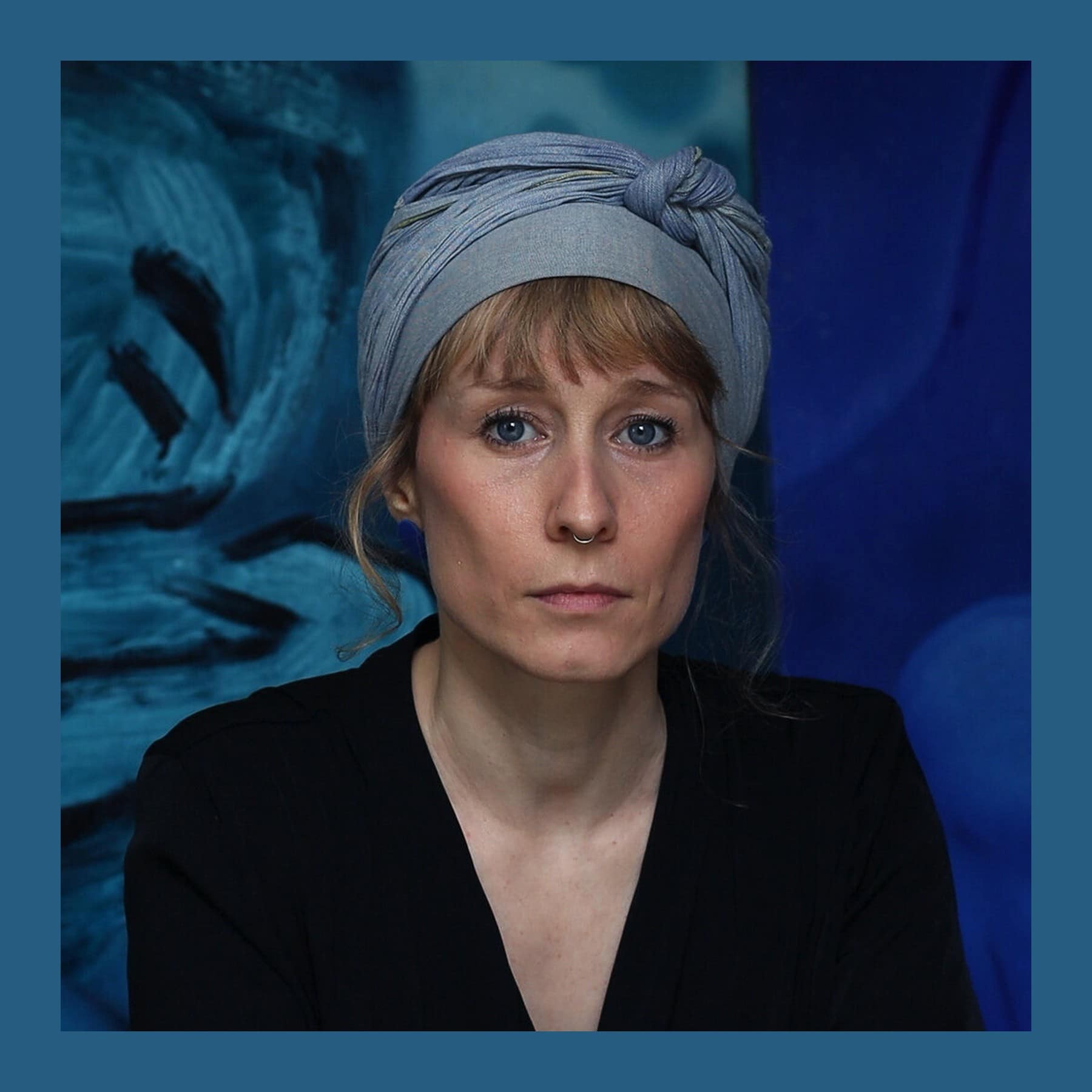 Aleah Chapin in a grey headscarf and black top in front of one of her blue paintings