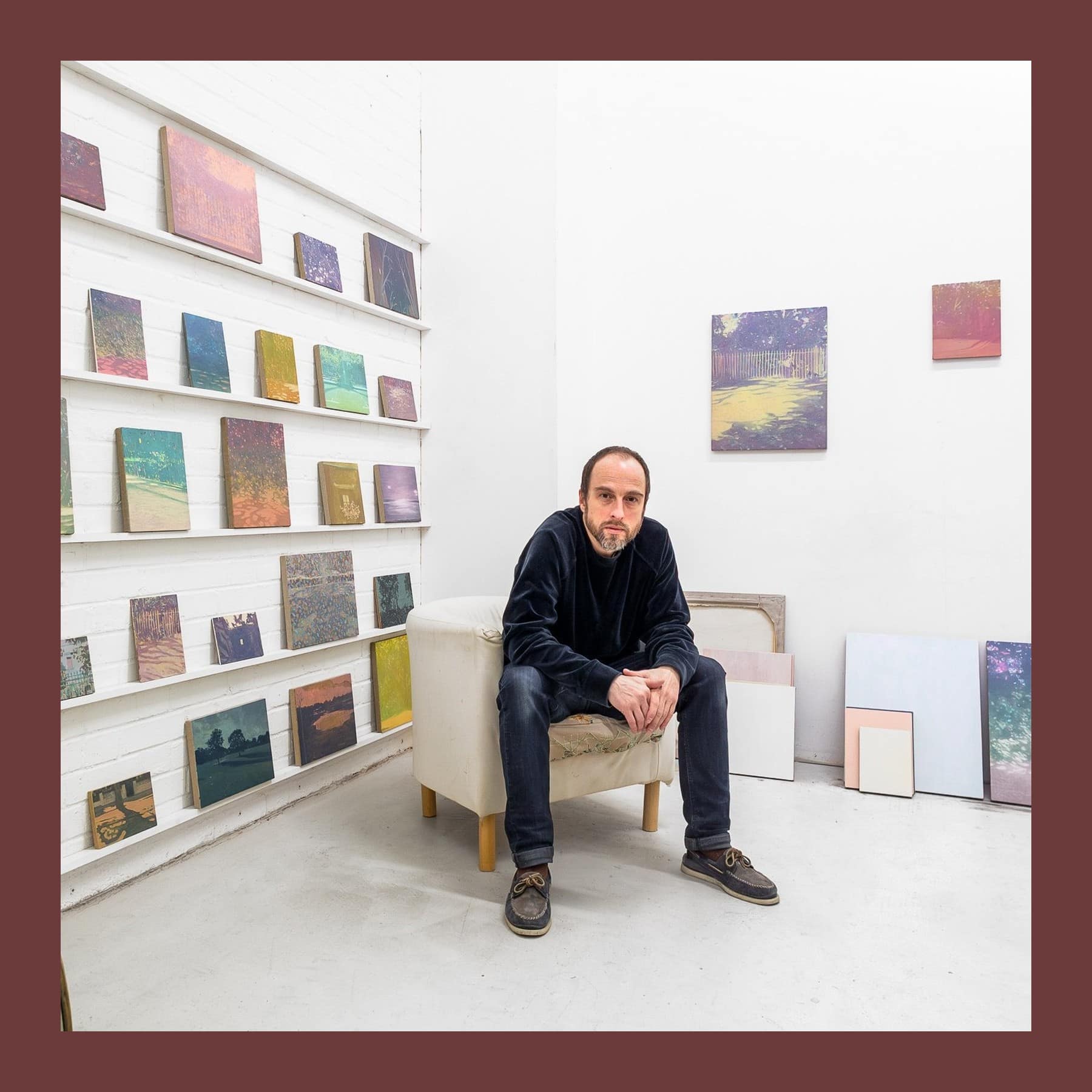 Salvatore Fiorello sitting in an armchair in his London studio, with rows of small landscape paintings on shelves beside him