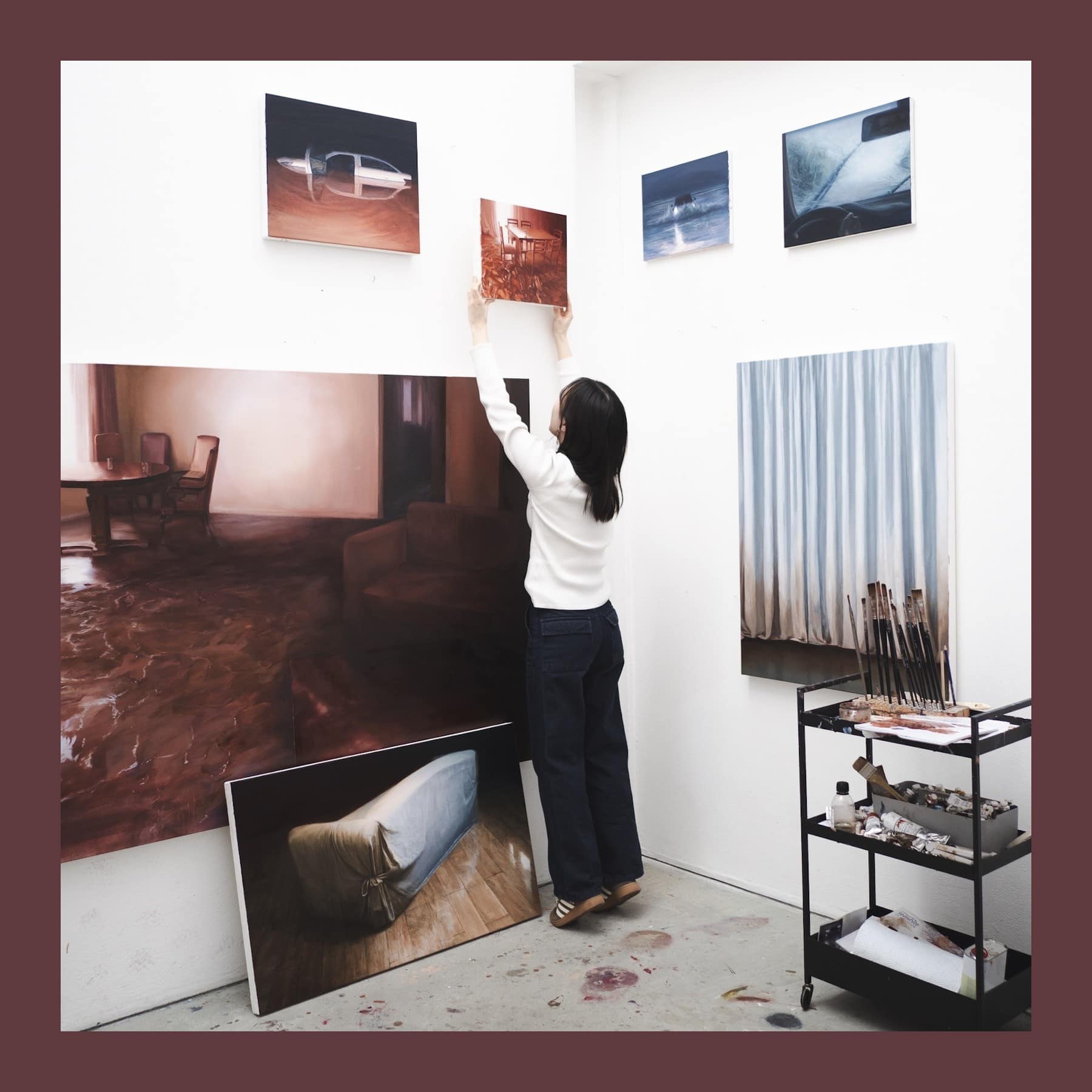 Sunkyung Baek hanging a small painting on her studio wall, surrounded by large paintings of flooded rooms and objects