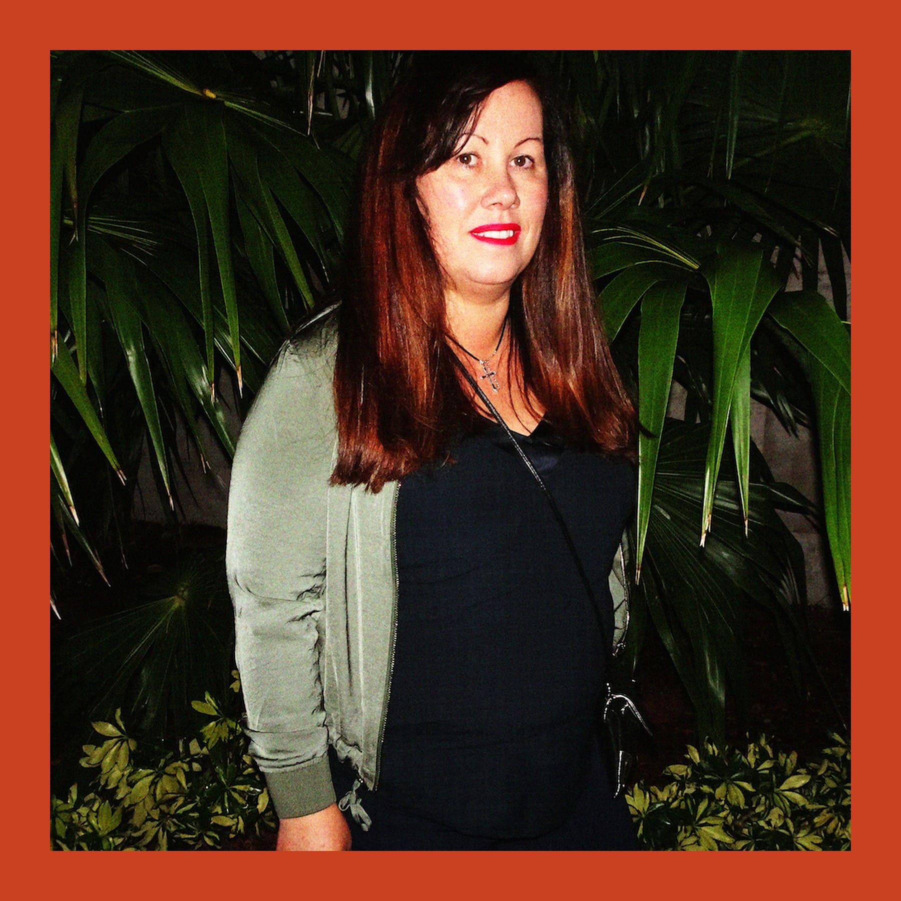 Genieve Figgis with long red hair, in a green cardigan, standing in front of palm leaves at night