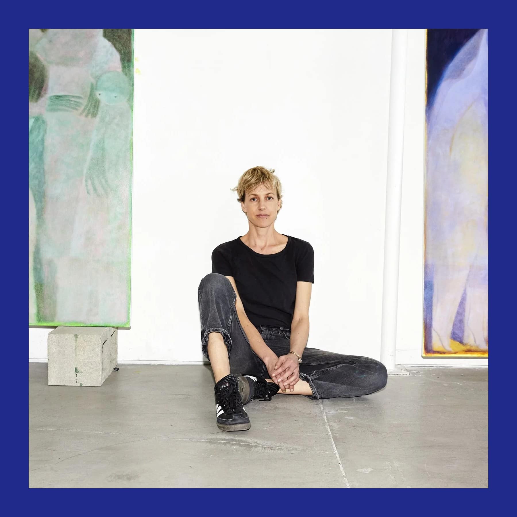Tahnee Lonsdale sitting cross-legged on a concrete studio floor between two large paintings of pale, ghostly figures