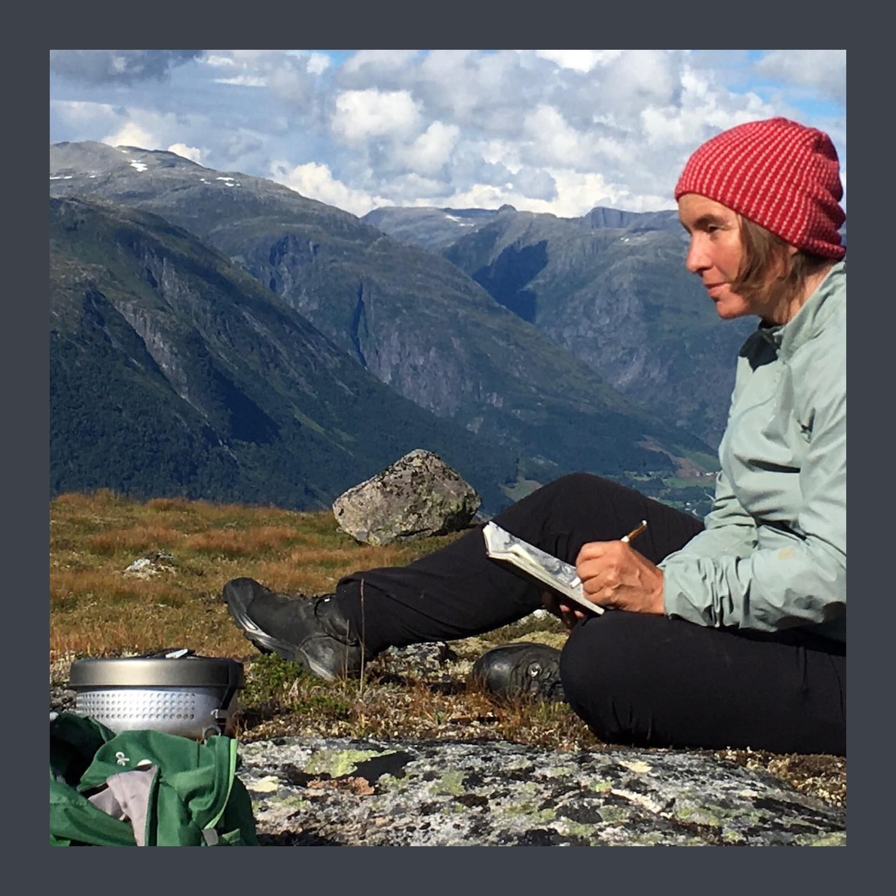 Heidrun Rathgeb in a red knitted hat sitting on a rocky mountainside, drawing in a sketchbook above a Norwegian valley