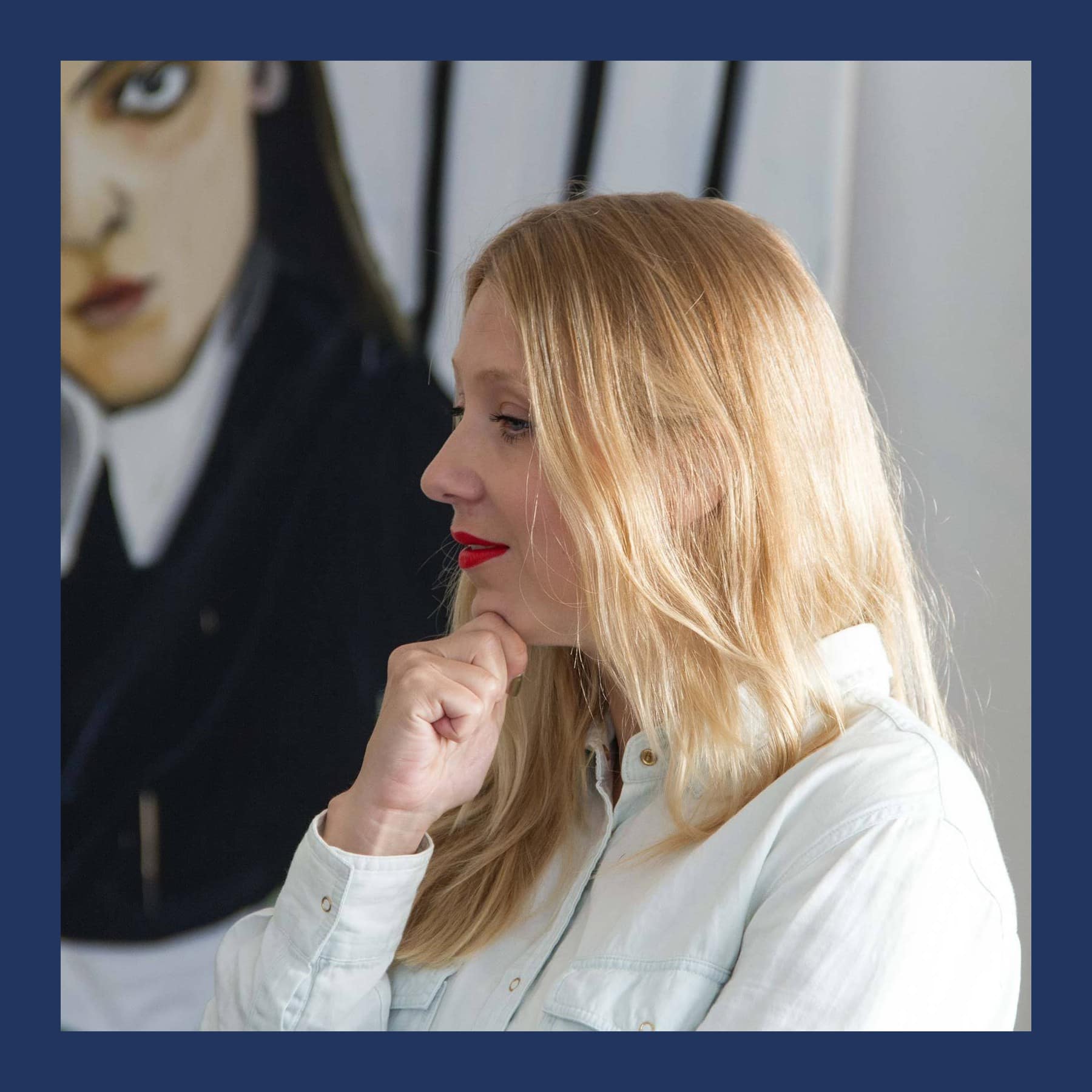 Jenni Hiltunen with long blonde hair and red lipstick, chin on her hand, in front of one of her large portrait paintings