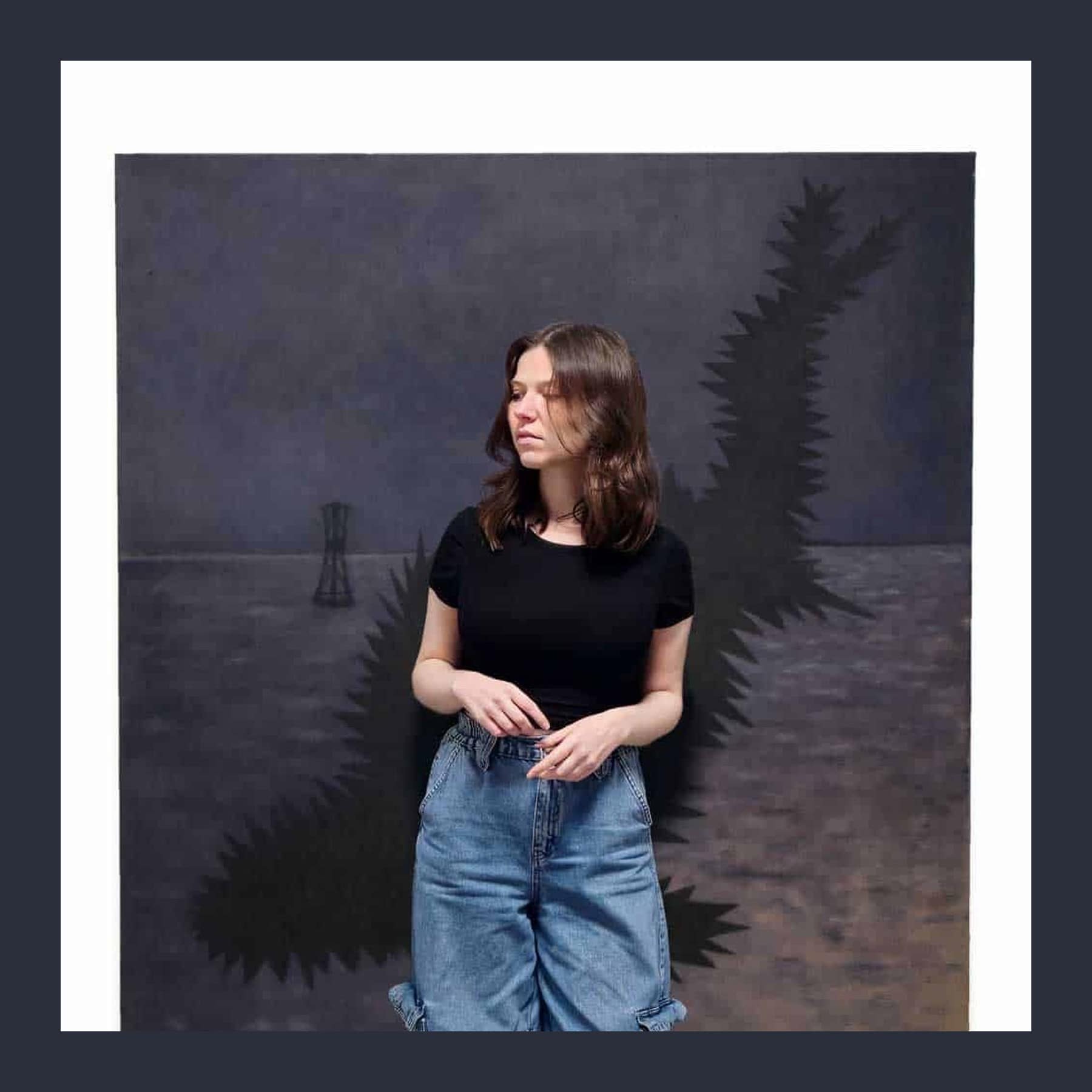 Grace Lee standing in front of one of their large dark paintings, wearing a black T-shirt and wide blue jeans