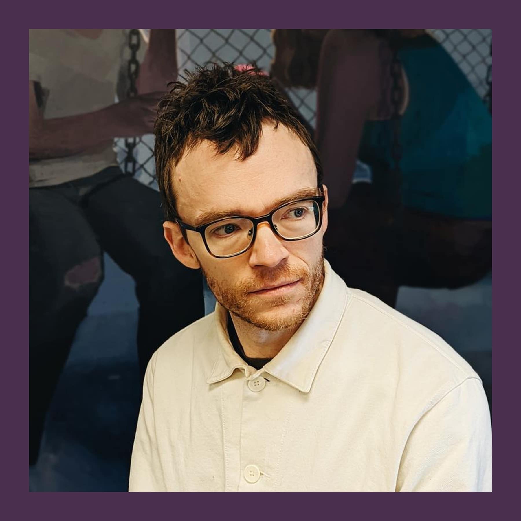 Matt Bollinger in glasses and a cream work jacket, sitting in front of one of his large paintings of figures by a chain-link fence