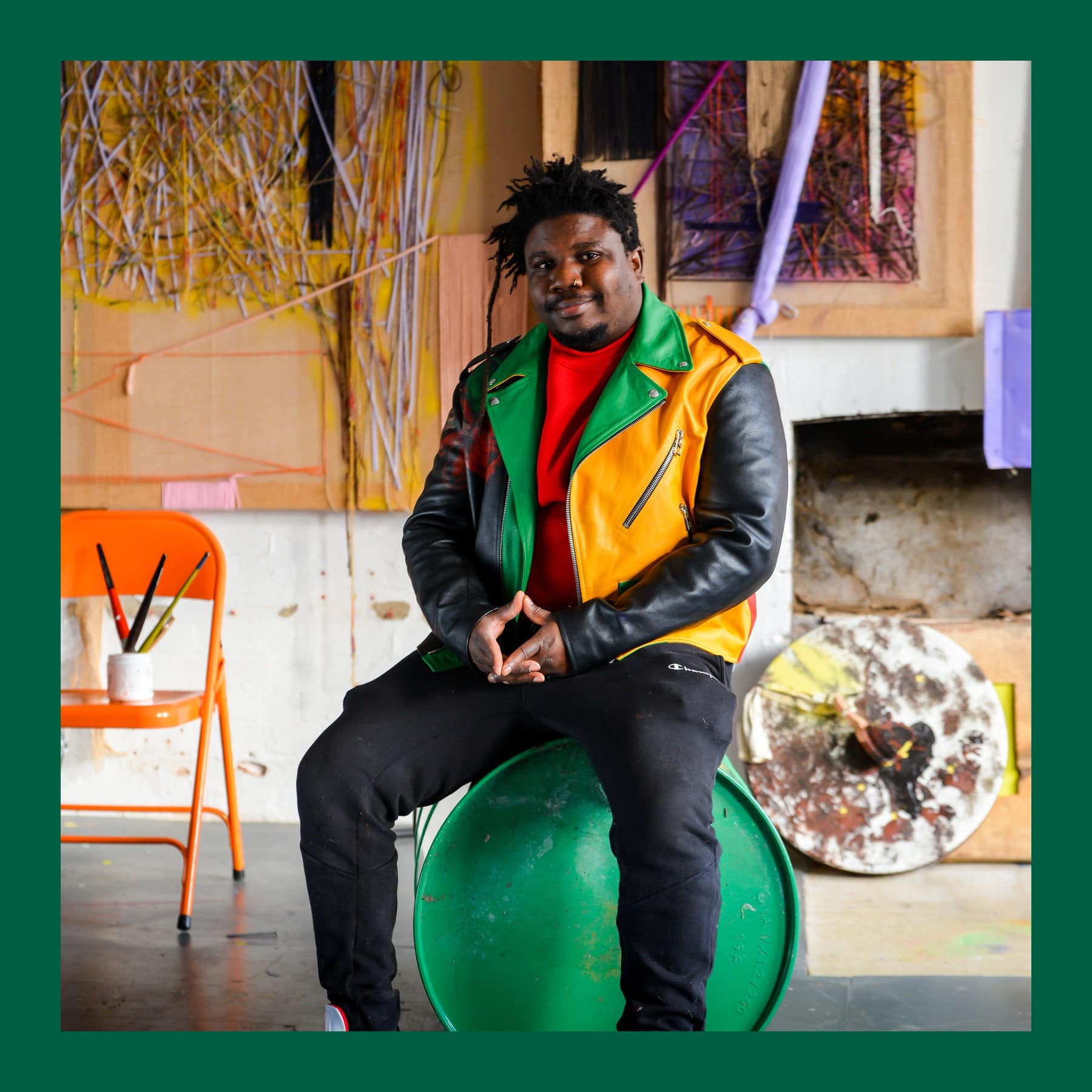 Alvaro Barrington in a green, yellow and black leather jacket, seated on a green drum in his studio