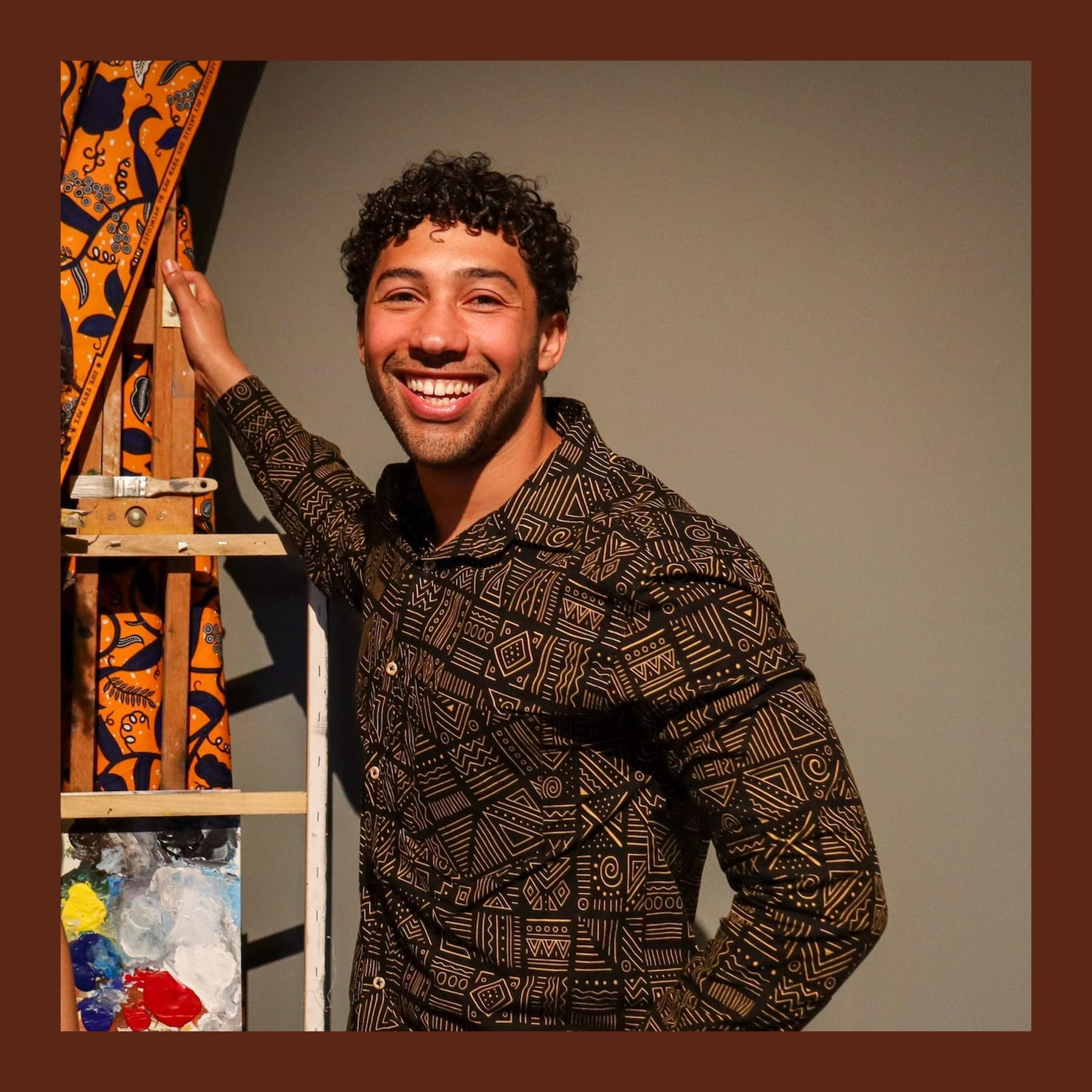 Joshua Donkor smiling, in a patterned brown shirt, with one hand on an easel draped in African wax-print fabric
