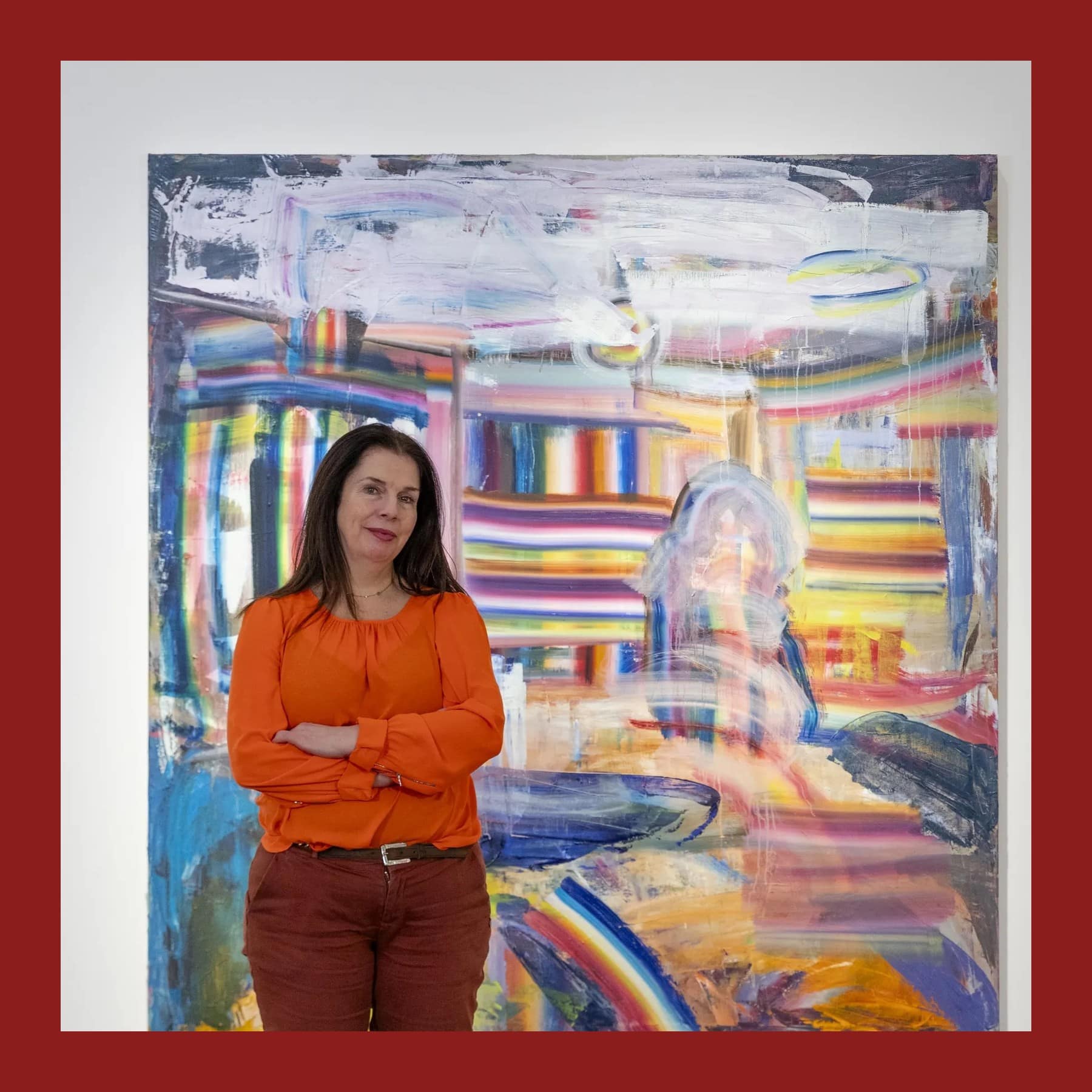 Diana Copperwhite in an orange blouse, arms folded, standing in front of one of her large abstract paintings with rainbow bands