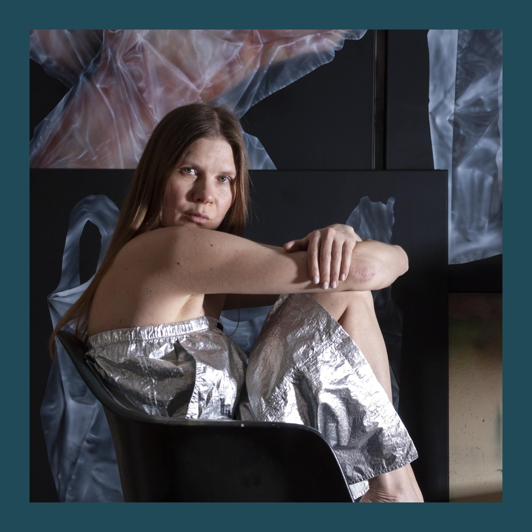 Helena Minginowicz sitting on a black chair in silver shorts, in front of her large airbrushed paintings of plastic bags