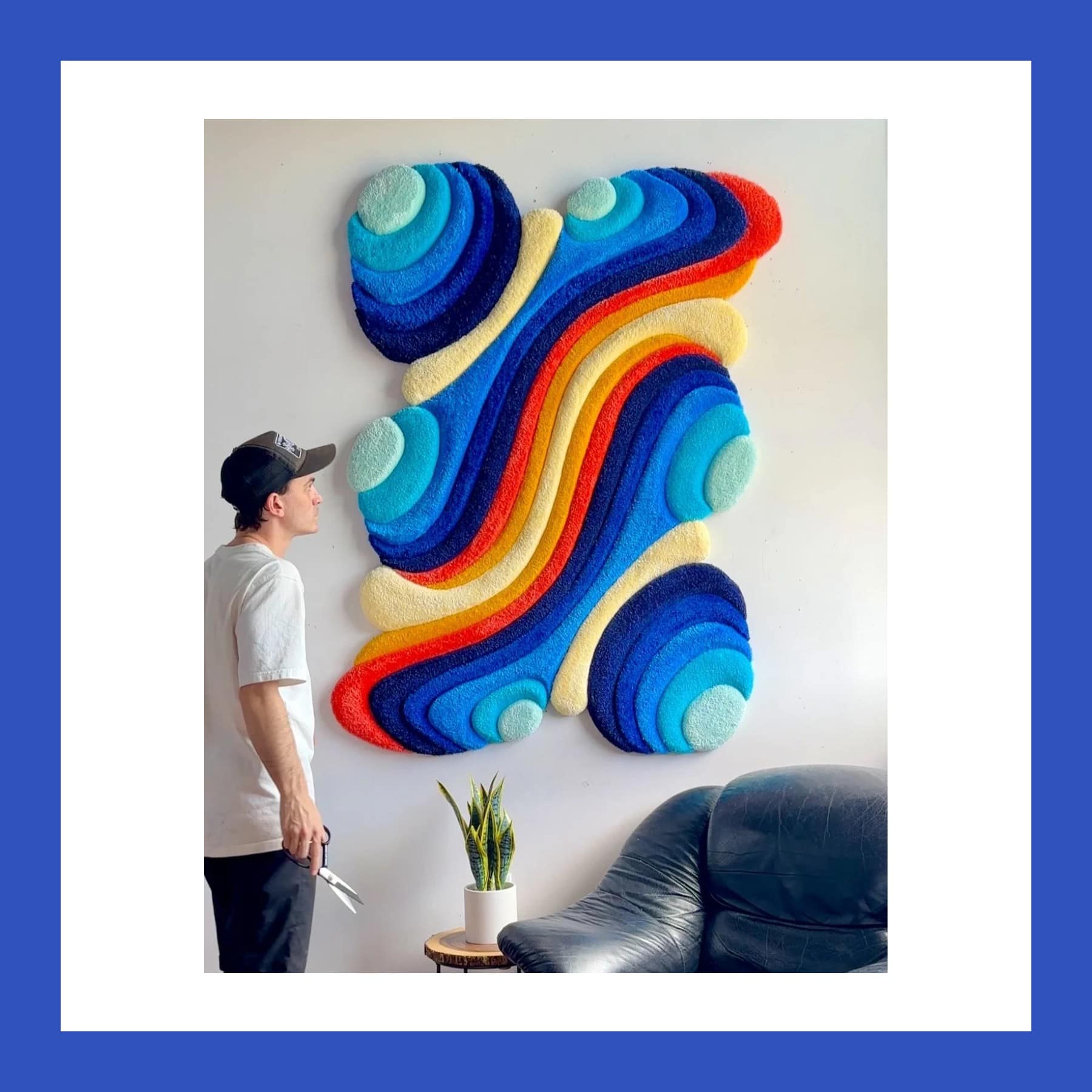 David Nott in a cap and white T-shirt, holding scissors, looking up at a large wavy tufted wall piece in blues, red and yellow