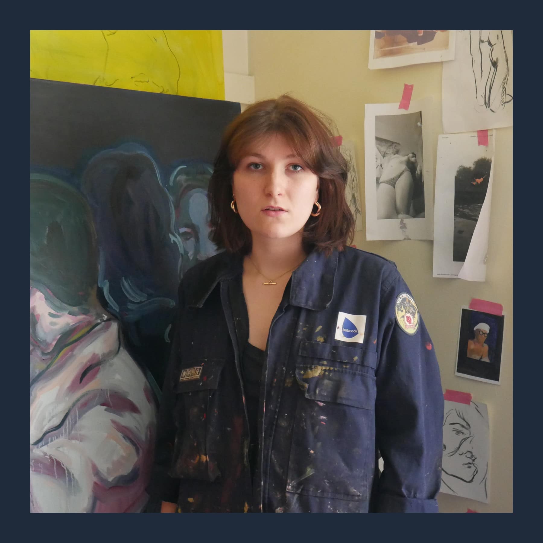 Abigail McGinley in a paint-spattered navy work jacket in her studio, beside one of her figure paintings and pinned-up drawings