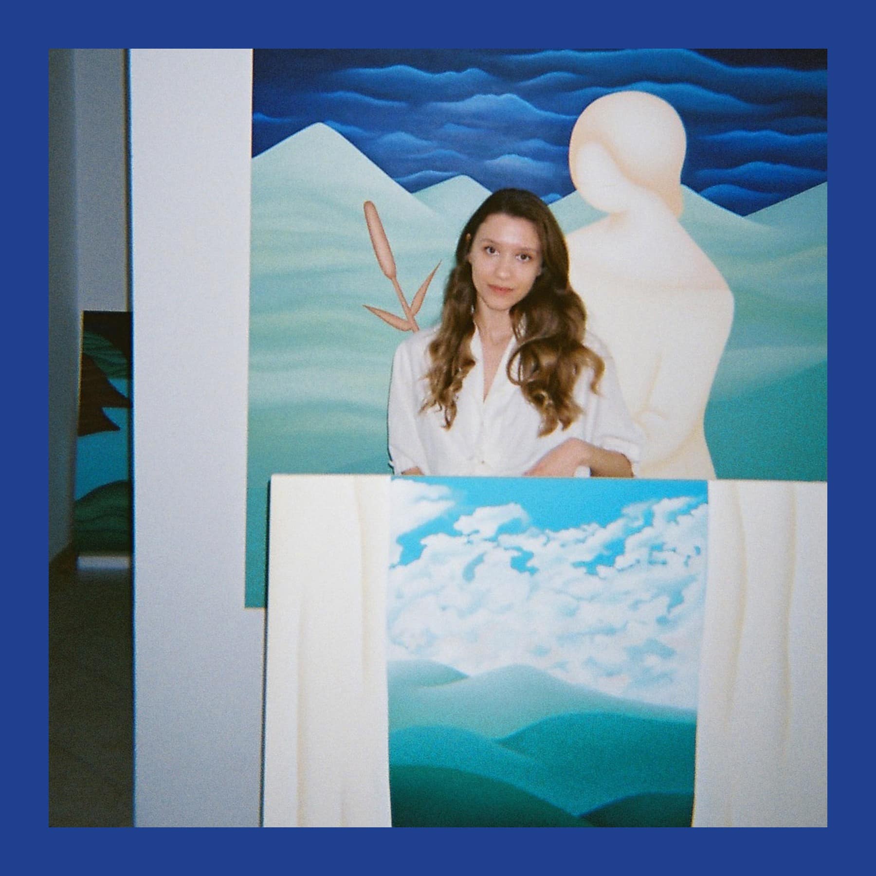 Anaja Hvastija Gaia in a white shirt standing among her paintings of pale figures, mountains and clouds