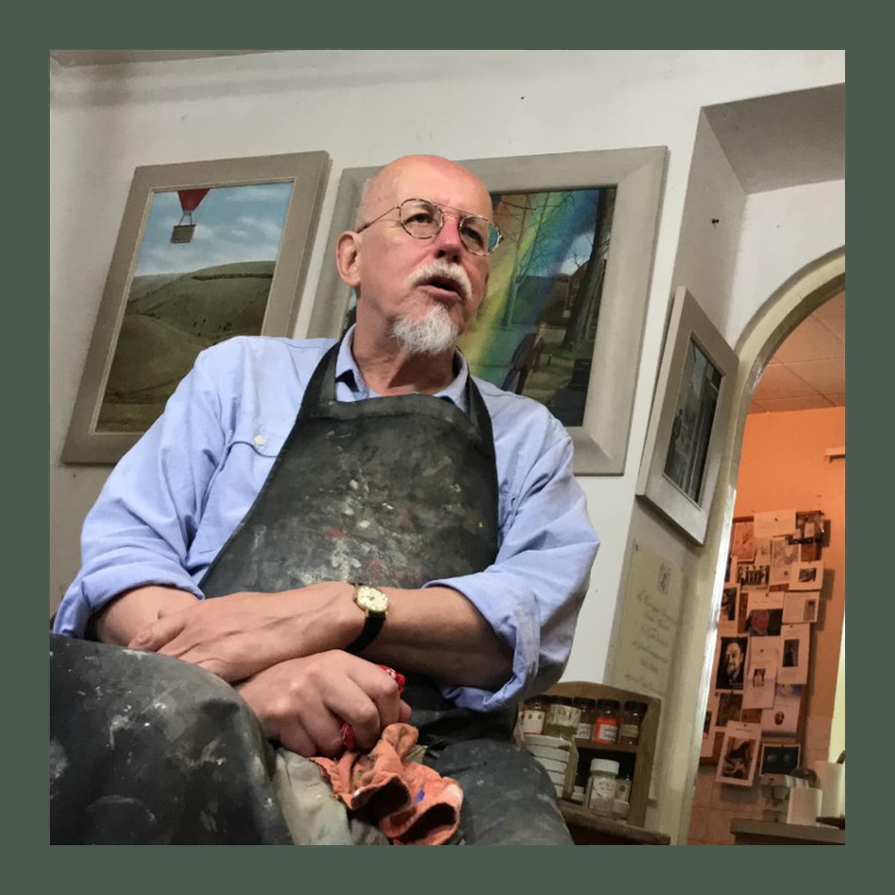 Peter Messer, with glasses and a white beard, in a paint-stained apron in his Lewes studio, surrounded by framed paintings