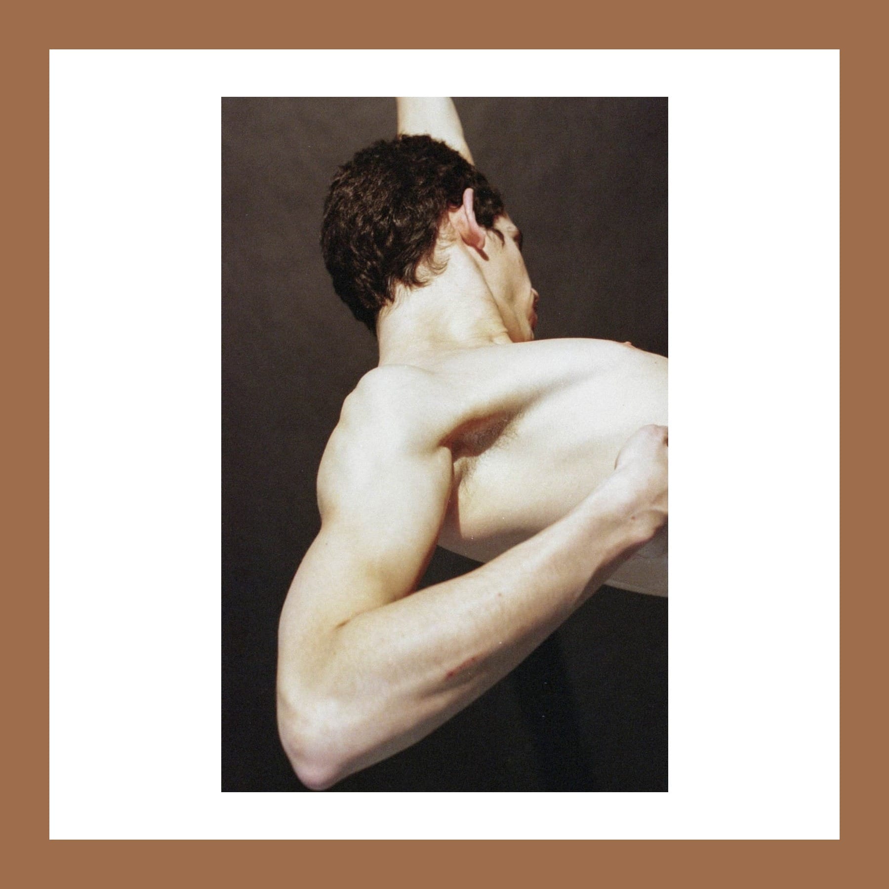 Colour photograph of a shirtless young man twisting, one arm wrapped tightly across his back and shoulder