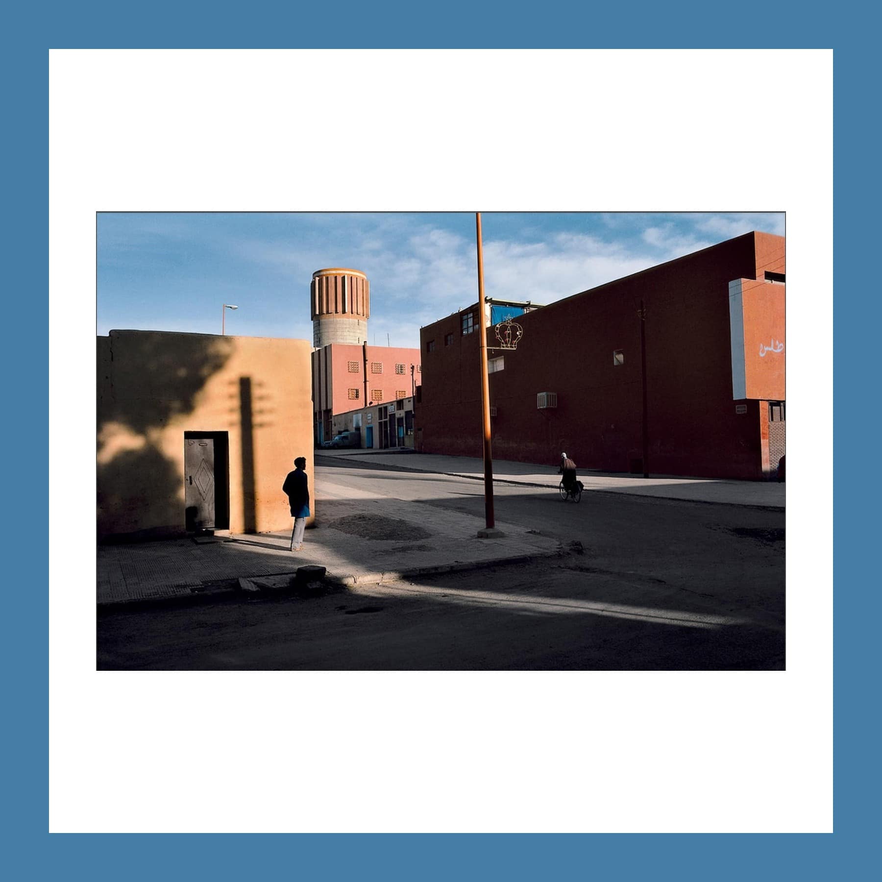 Colour photograph of a sunlit Moroccan street corner with red and ochre walls, a water tower and a man standing in shadow