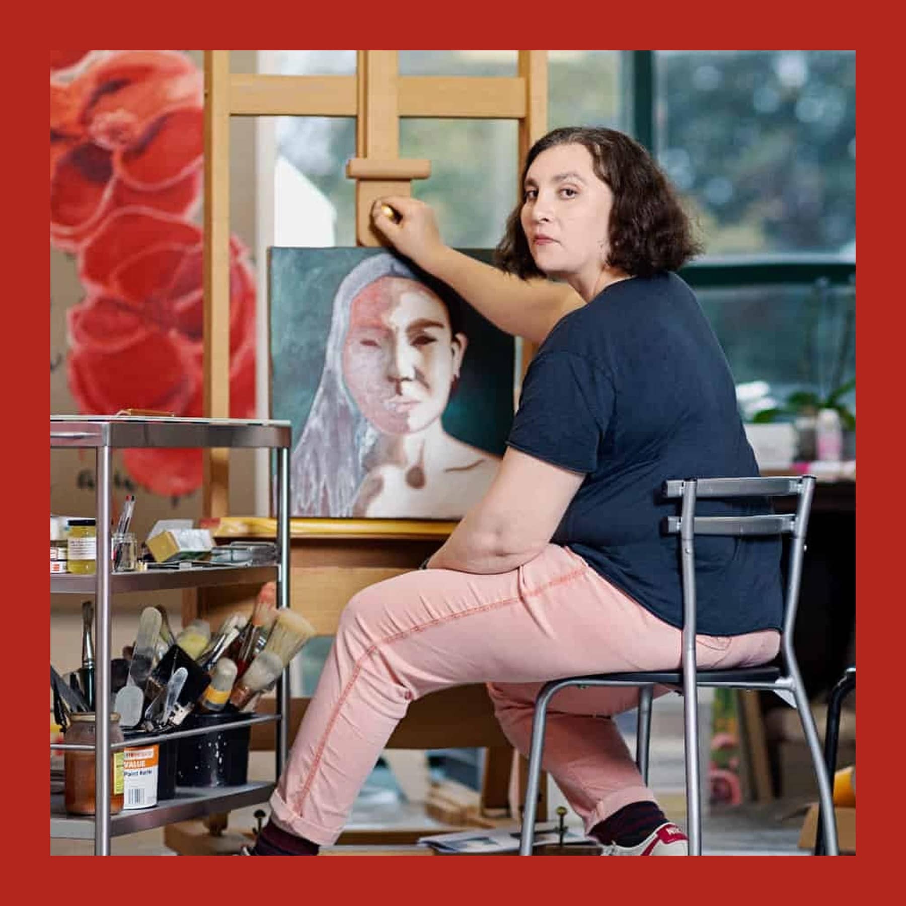 Victoria Cantons sitting on a metal chair at her easel, with one hand on an unfinished portrait, in a studio full of paint and brushes