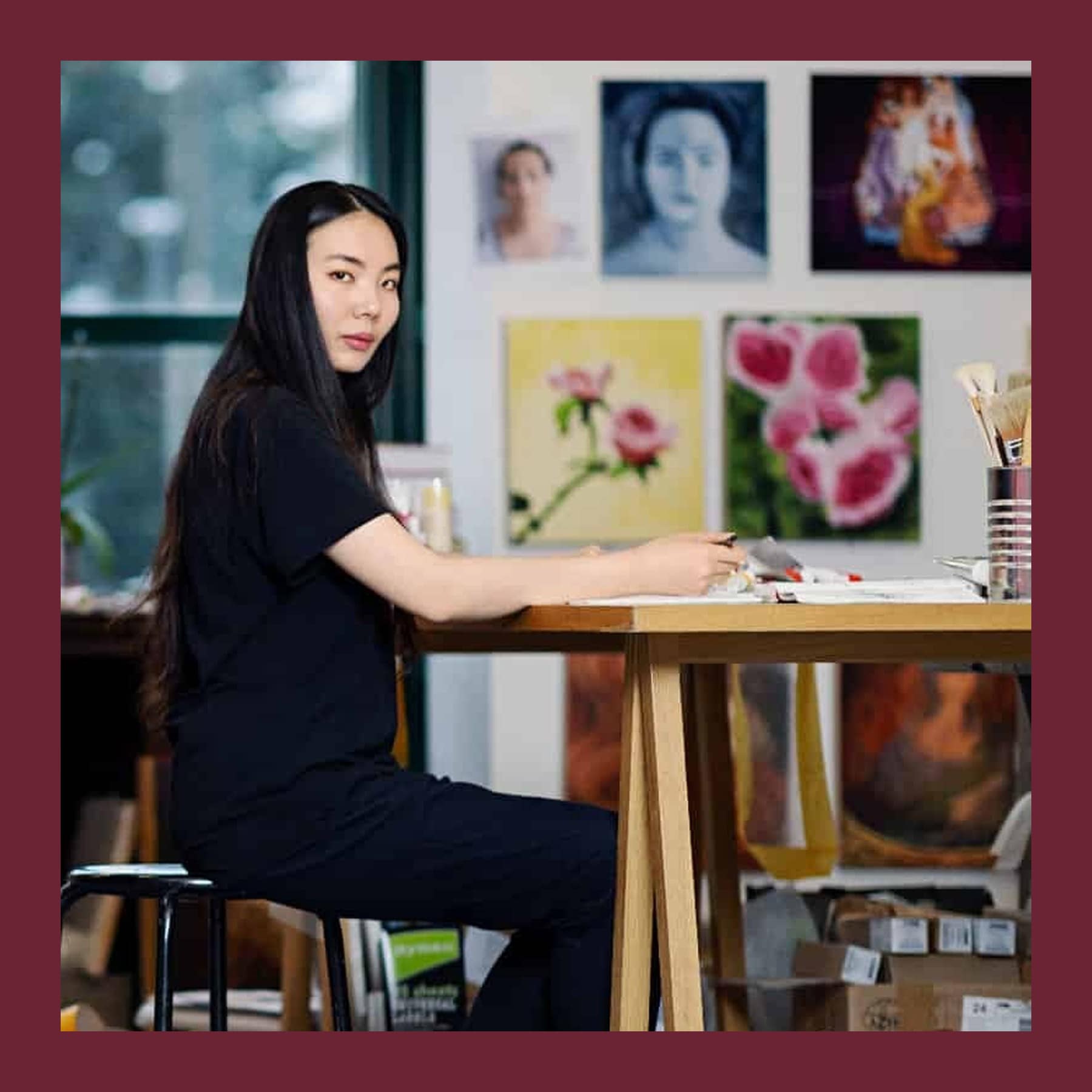Xu Yang sitting at a wooden studio table with a pencil in hand, in front of a wall of her paintings of roses and faces