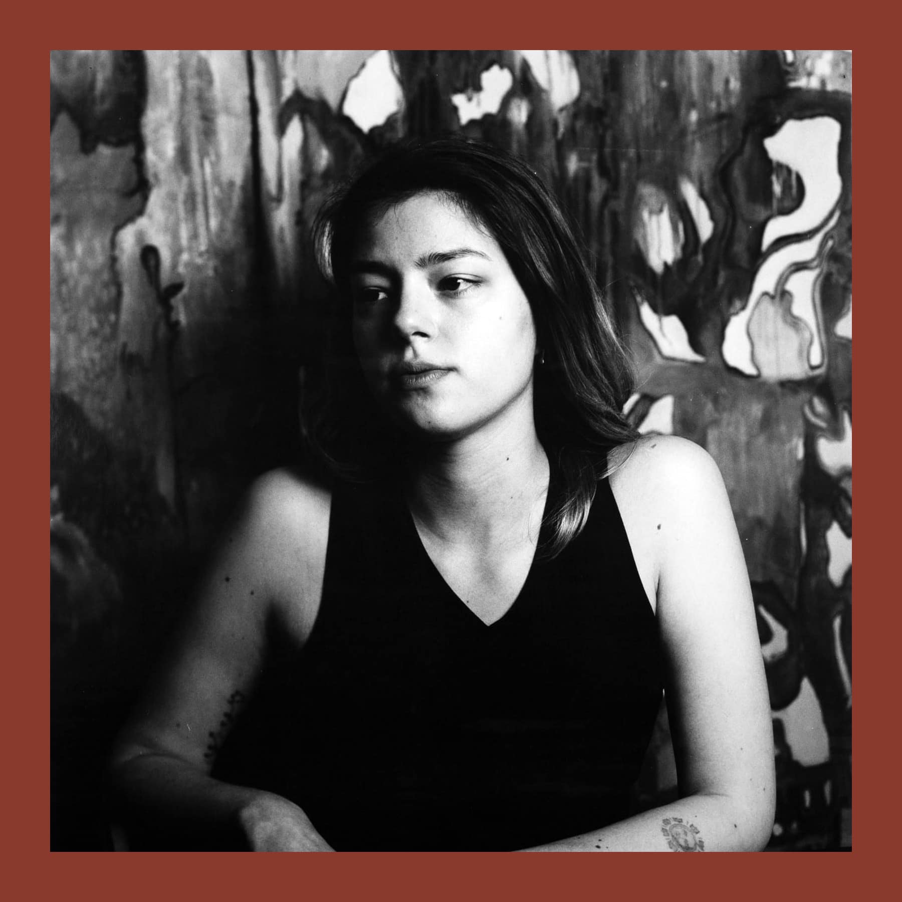 Black and white photograph of Alexandra Beteeva in a dark vest top, seated in front of a large abstract painting