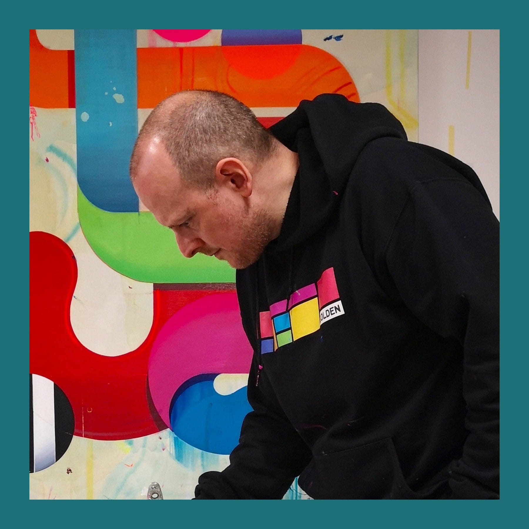 Nick Grindrod in a black hoodie leaning over to work in front of one of his large abstract paintings of looping red, green and blue forms