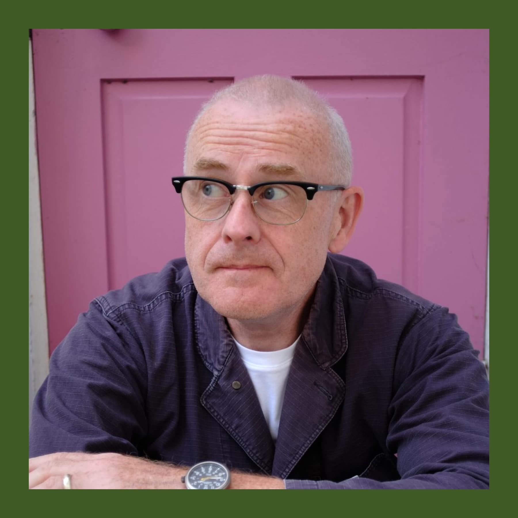 Colin Murphy in black-rimmed glasses and a navy work jacket, arms folded, in front of a pink door
