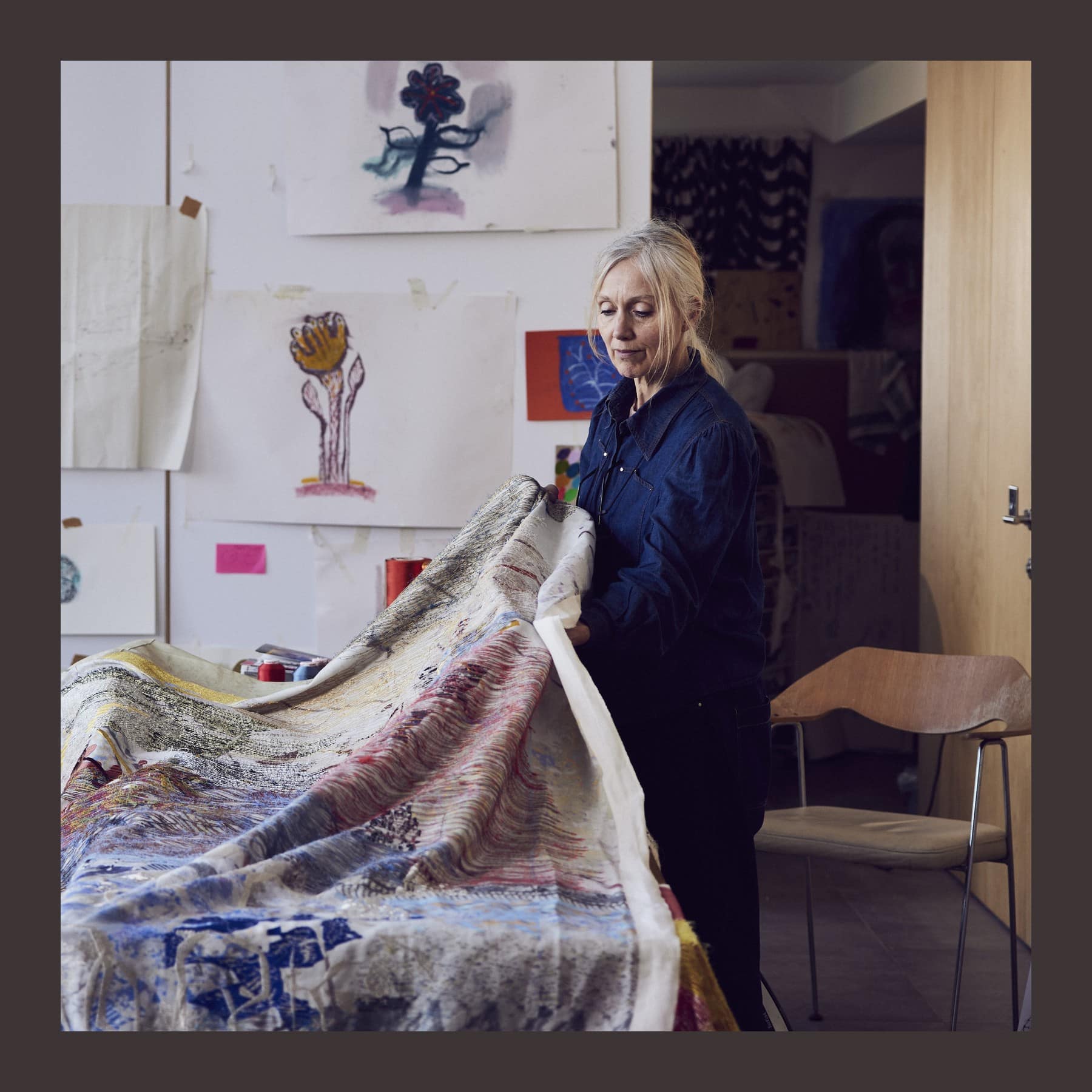Alice Kettle in a navy shirt in her studio, lifting a large stitched textile across a table, with drawings pinned to the wall behind