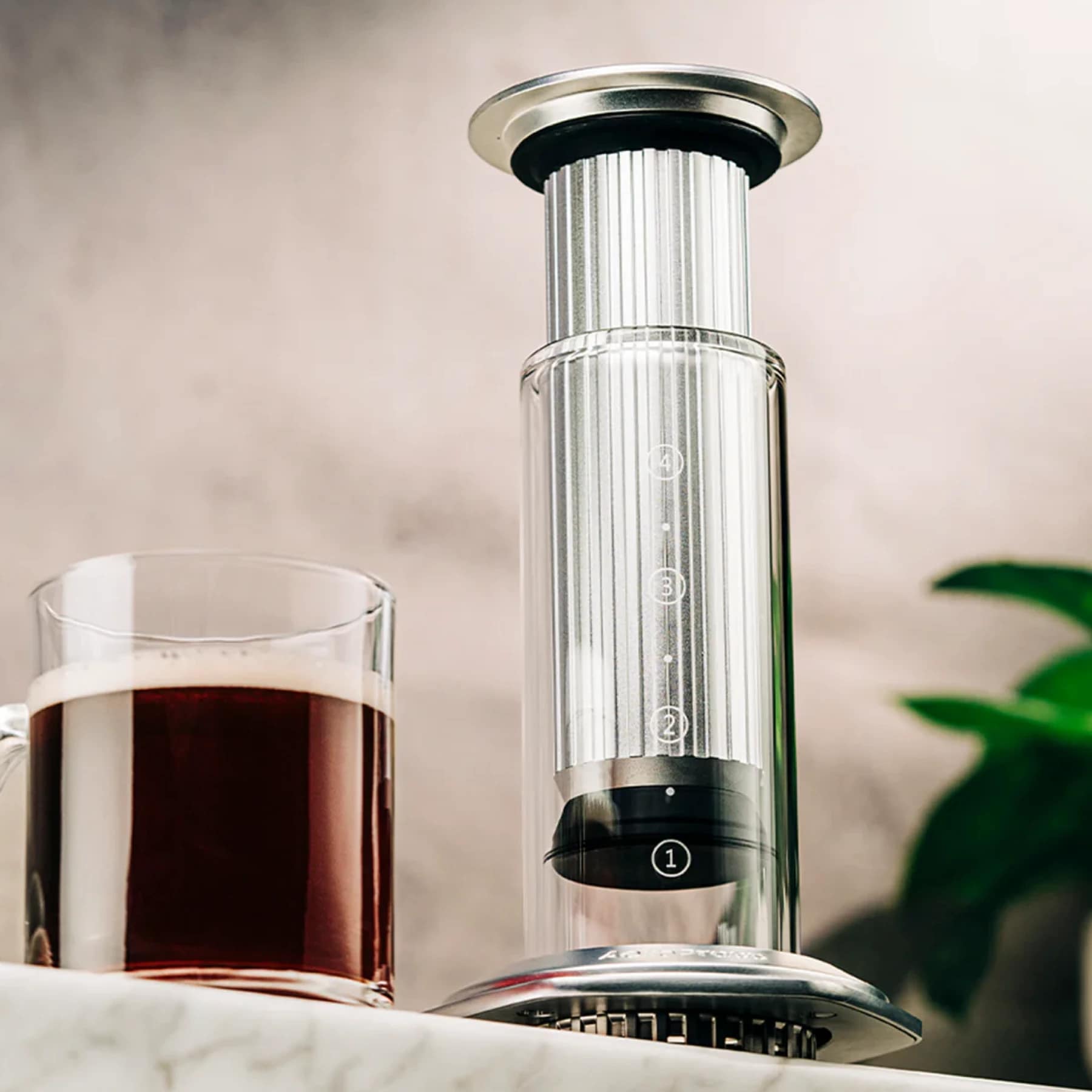 AeroPress Premium Coffee Maker with stainless-steel design and glass carafe beside a freshly brewed cup of coffee on a marble countertop.