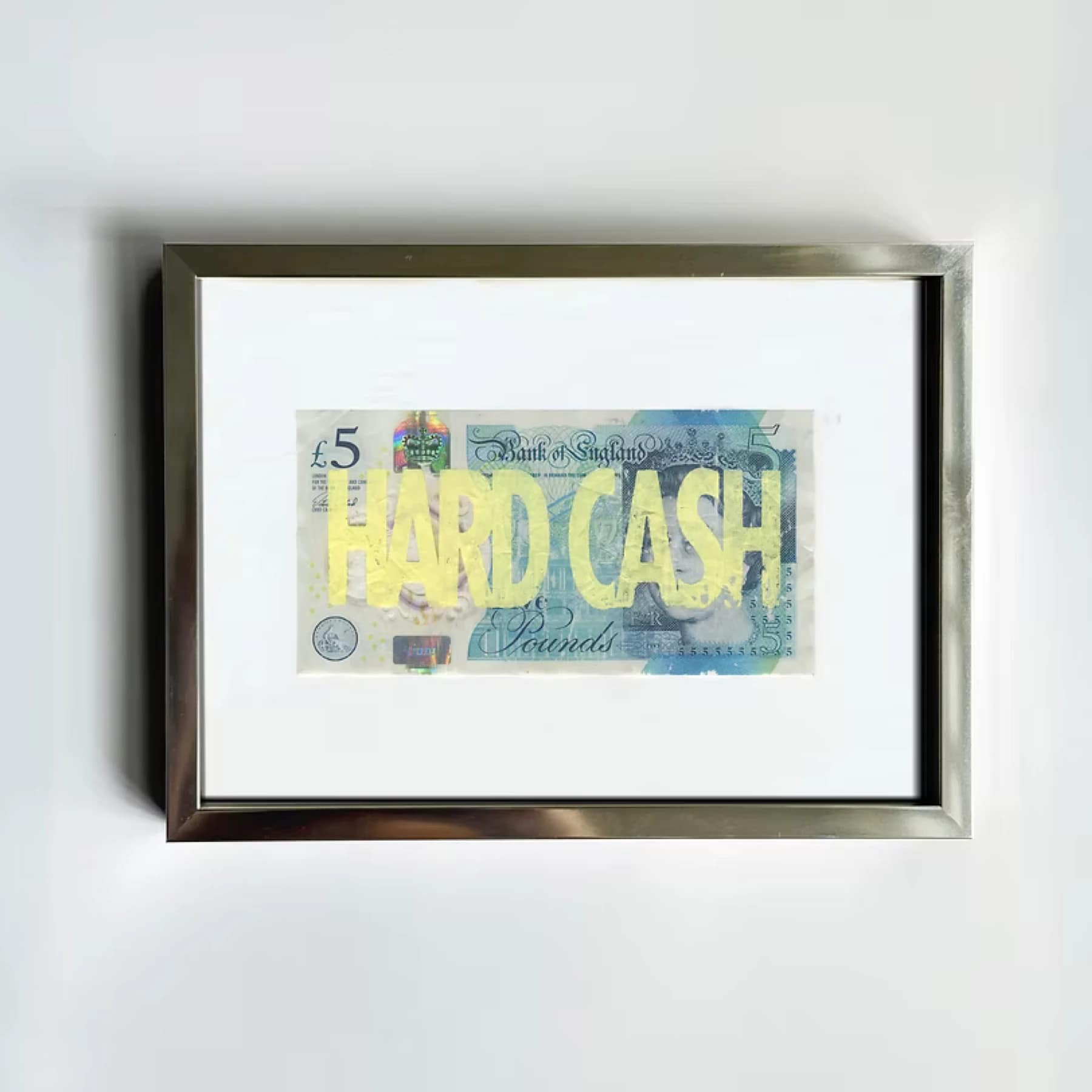 Framed Corbin Shaw Hard Cash print featuring bold yellow riso text over a £5 banknote, in a silver frame with white mount, hung on a white wall.