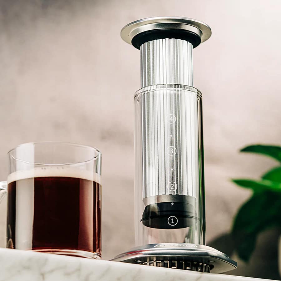 AeroPress Premium Coffee Maker with stainless-steel design and glass carafe beside a freshly brewed cup of coffee on a marble countertop.