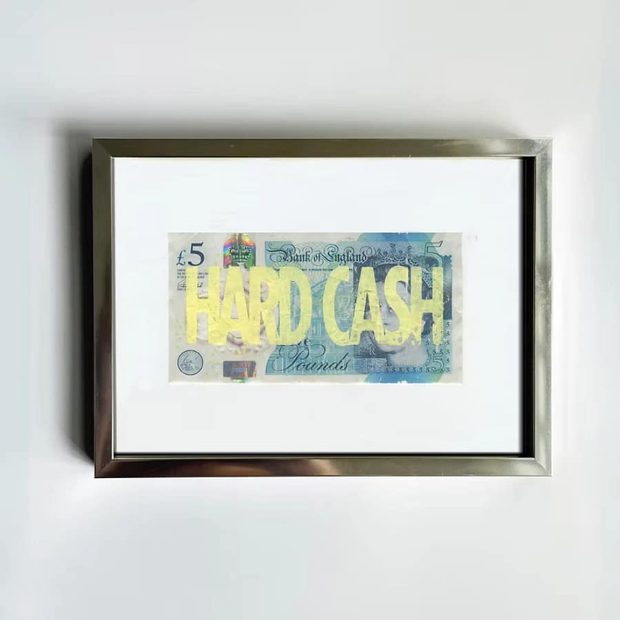 Framed Corbin Shaw Hard Cash print featuring bold yellow riso text over a £5 banknote, in a silver frame with white mount, hung on a white wall.