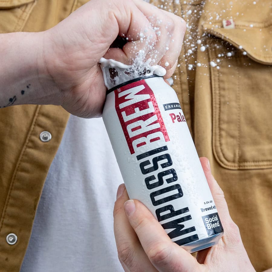 Hand opening a cold can of Impossibrew enhanced pale ale, with foam spraying, highlighting the refreshing alcohol-free Social Blend drink
