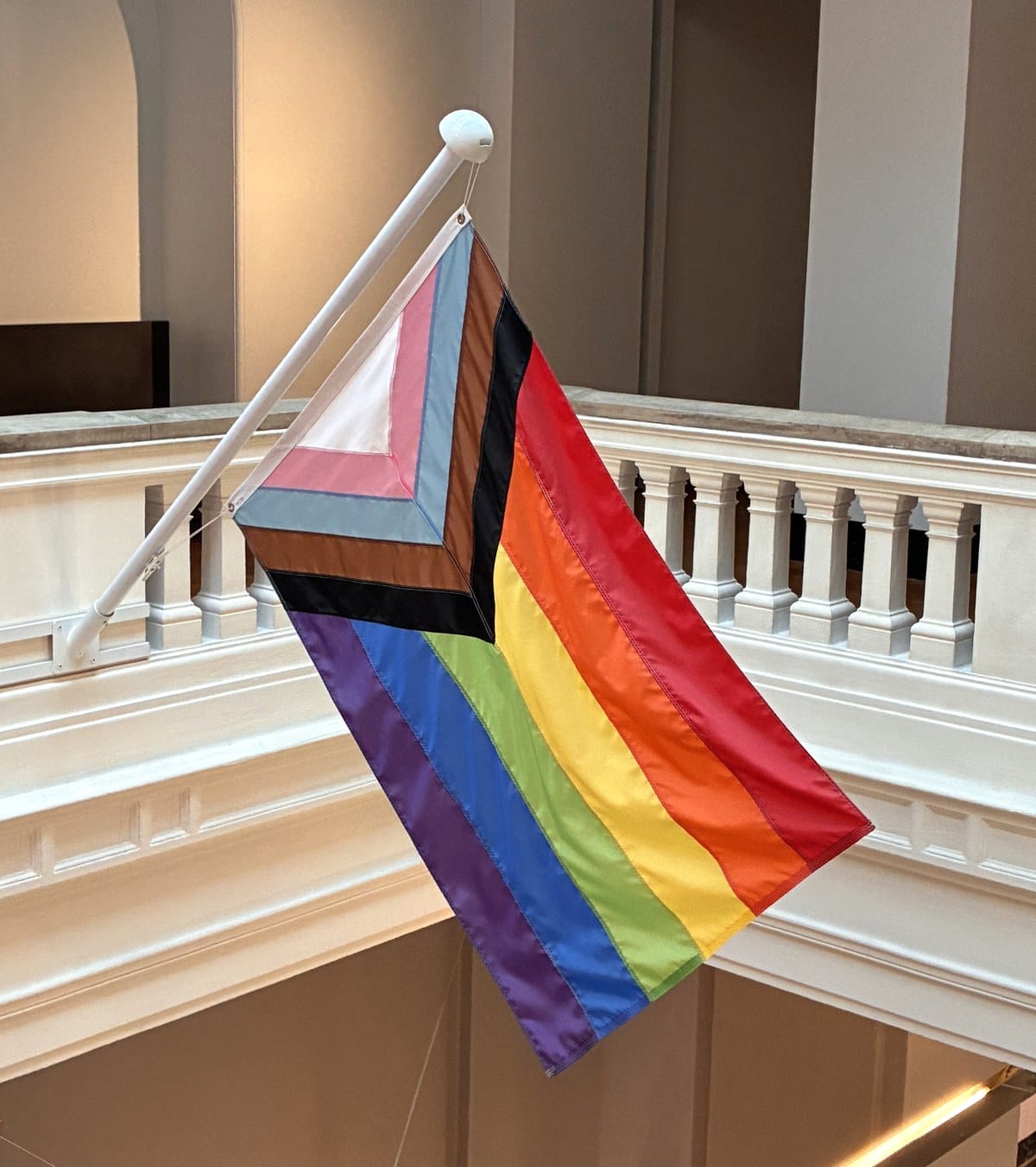 Progress Pride flag seen from above, flying over the marble sculpture corridor at the V&A